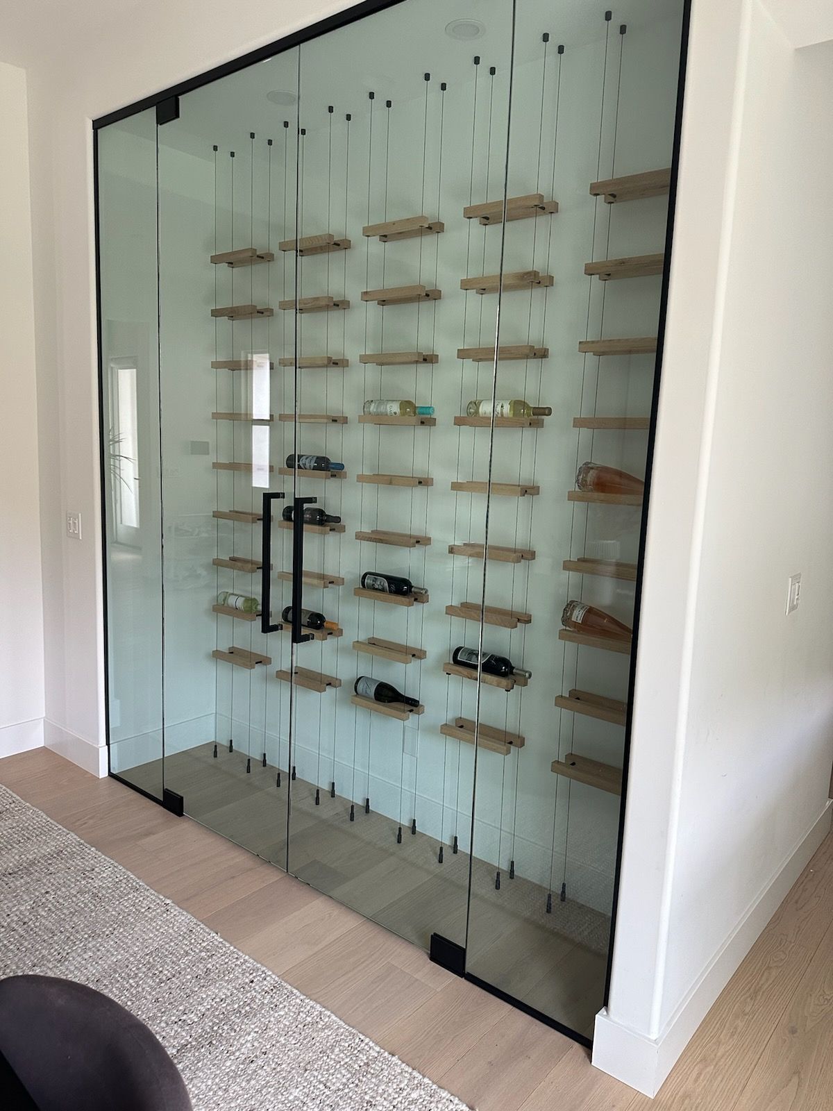 Glass-enclosed wine cellar with wooden shelves holding bottles. Black door, modern design, light wooden floor.