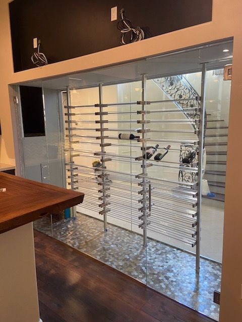 Wine cellar with glass walls, stainless steel racks, and bottles on display. Dark wood floors and a staircase are visible.