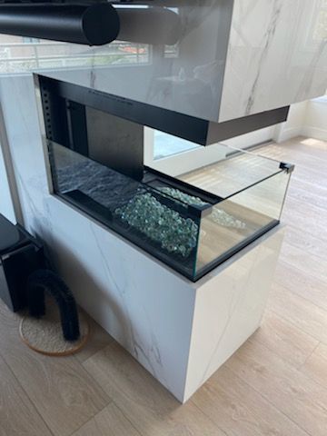 Modern kitchen island with glass-enclosed feature filled with decorative green glass. White marble paneling, wooden floors.