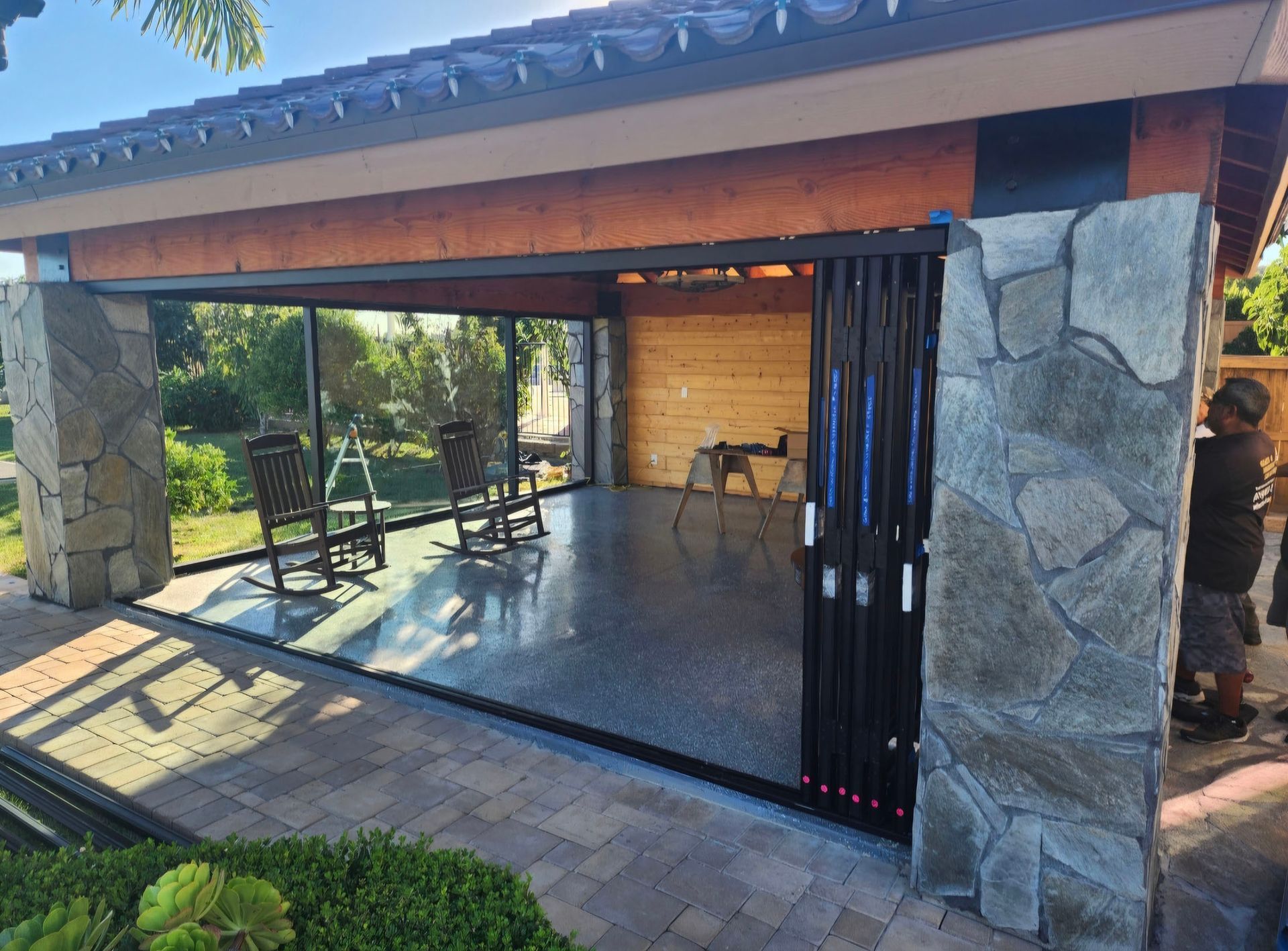 Outdoor patio with glass walls, stone columns, wooden roof, and closed folding doors; person to the right.