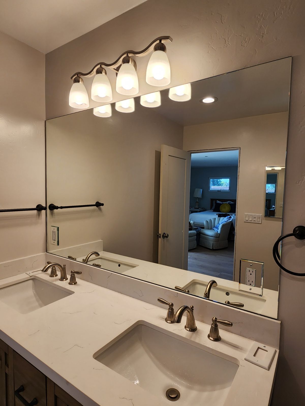 Bathroom with a large mirror, dual sinks, and a light fixture over the mirror. The door to the bedroom is open.