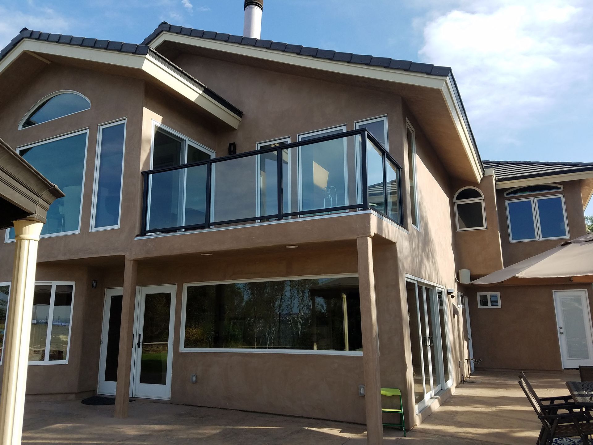 Two-story tan house with a glass balcony, large windows, and a sunny outdoor patio.