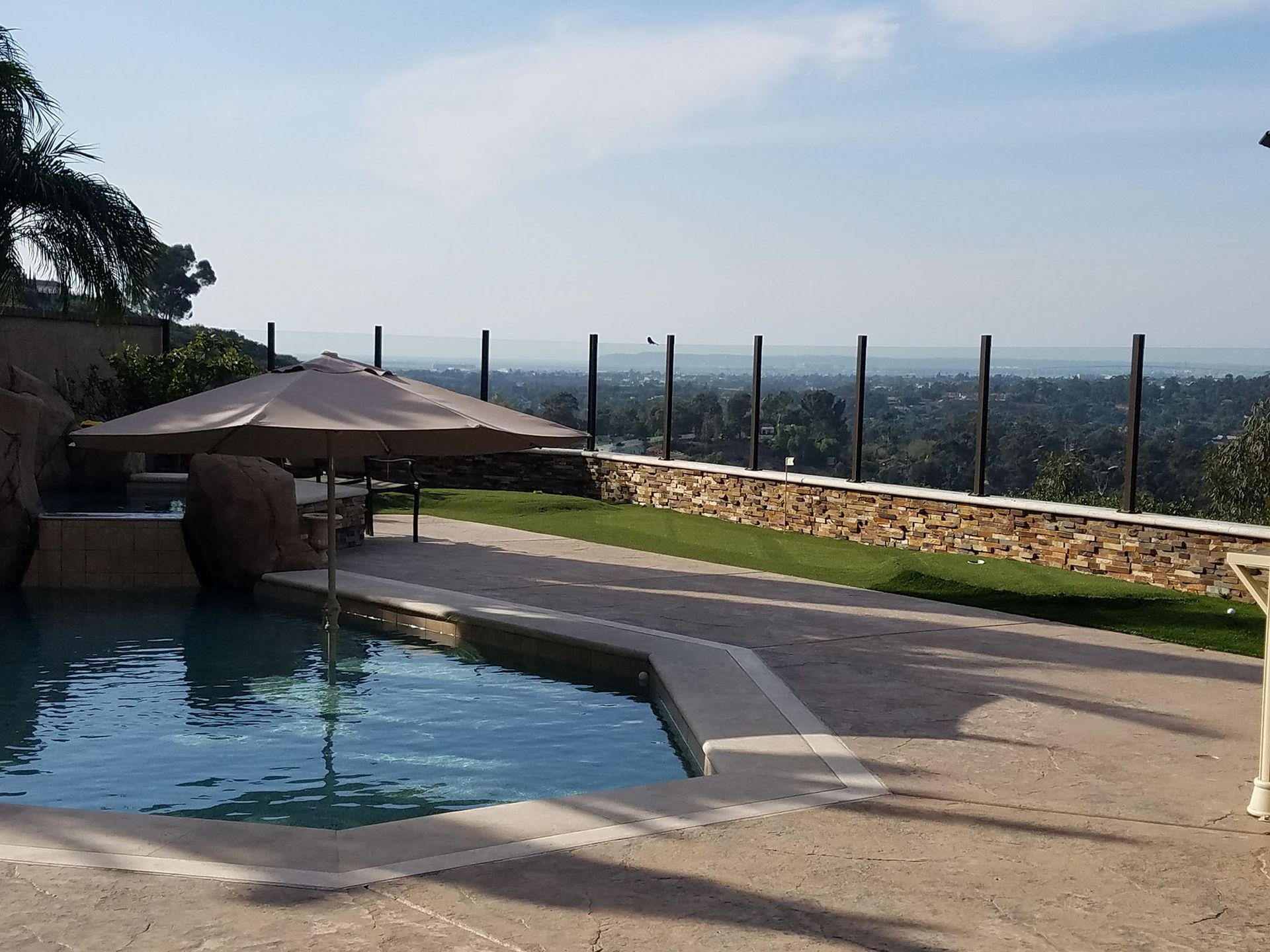 Poolside view with umbrella, pool, stone wall, and distant cityscape.