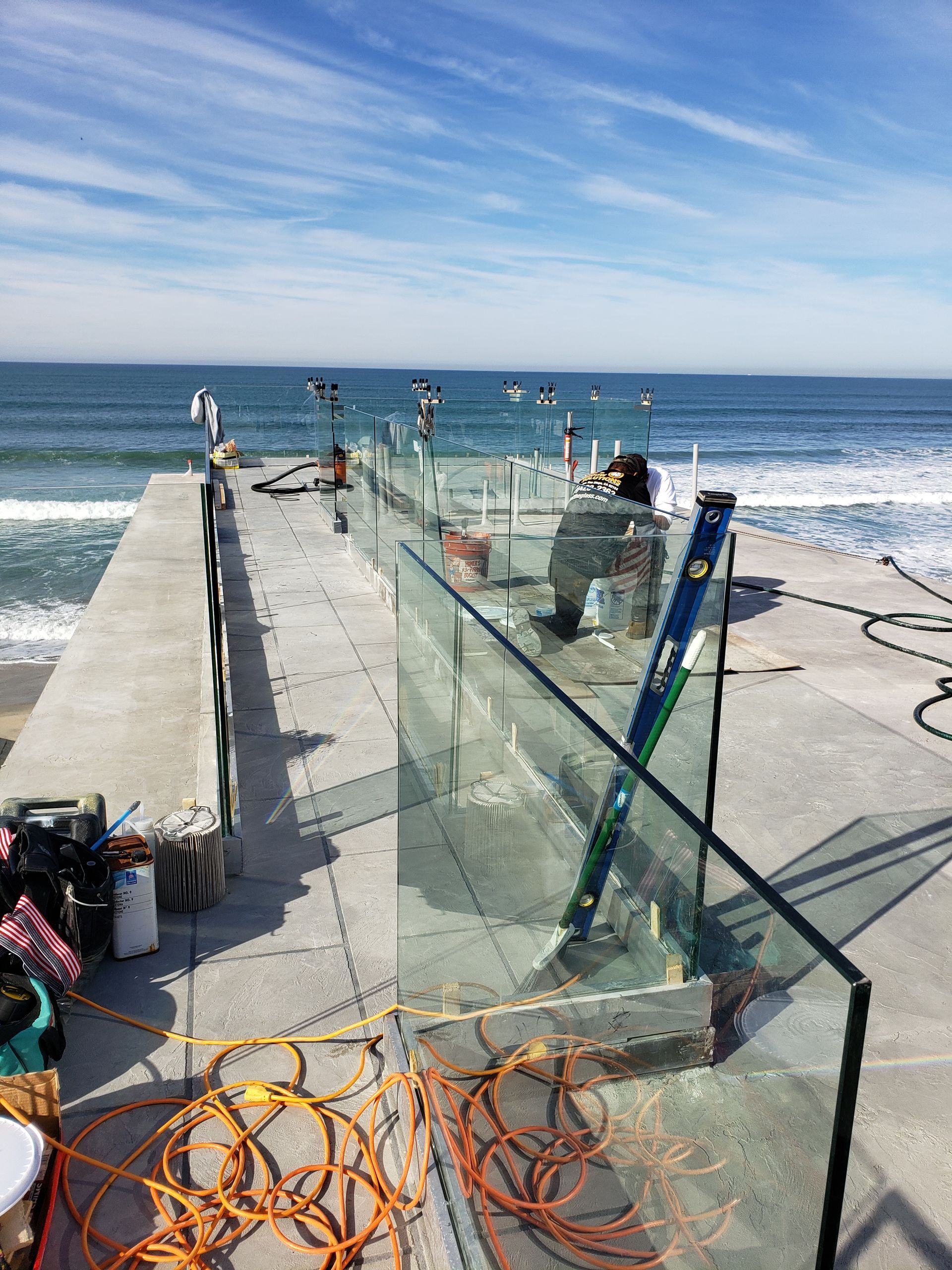 Construction of a glass barrier on a concrete pier overlooking the ocean. A worker uses a level. Blue sky.