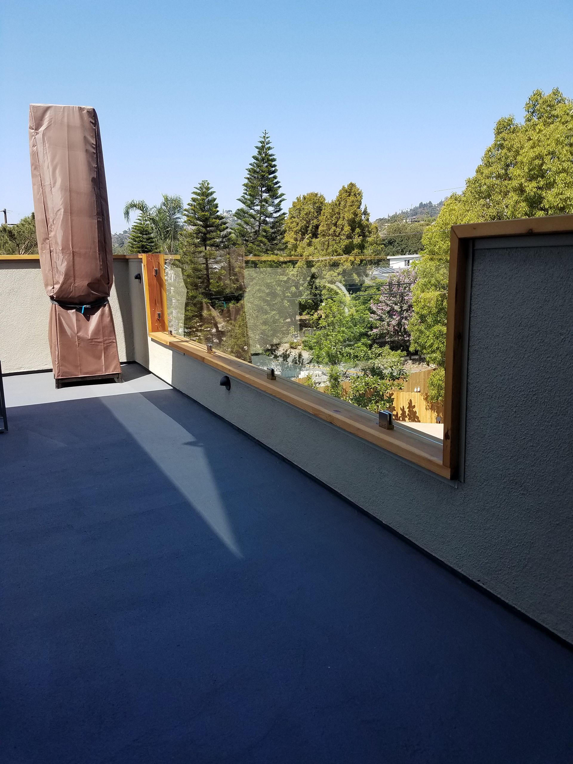 Rooftop deck with blue floor, textured gray walls, wooden railing, and glass panels overlooking a green landscape.
