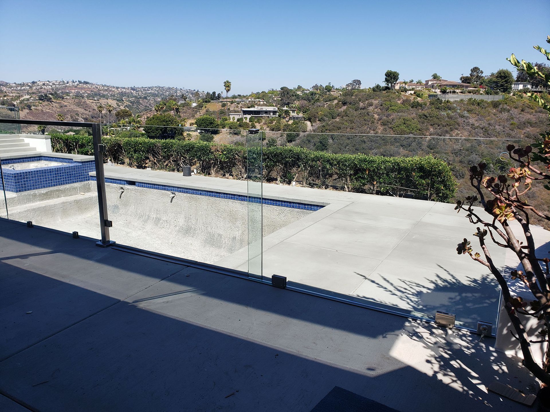 Empty pool with glass fencing, overlooking a hilly landscape on a sunny day.