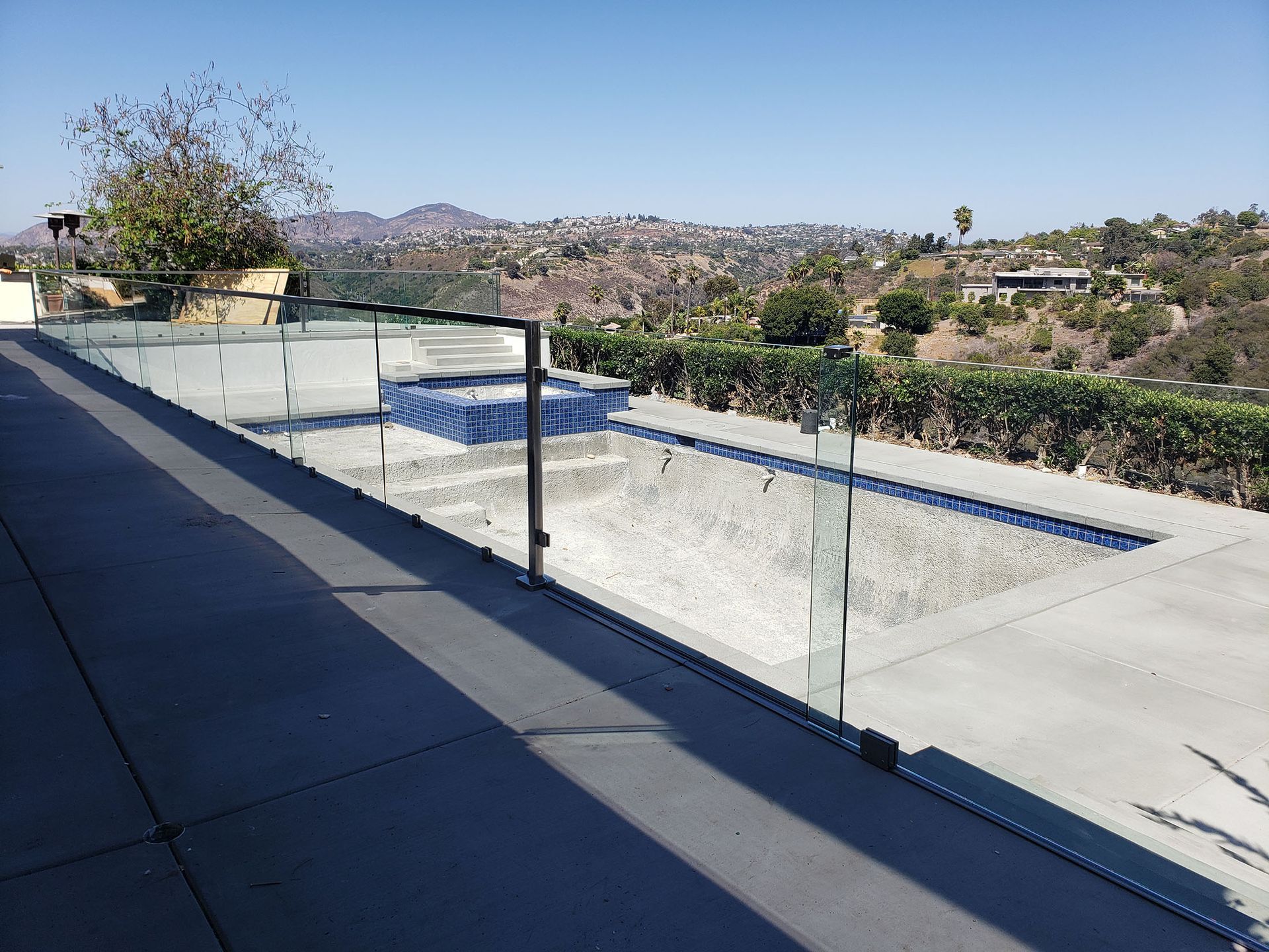 A glass fence surrounds an empty swimming pool. View of hills in the background.