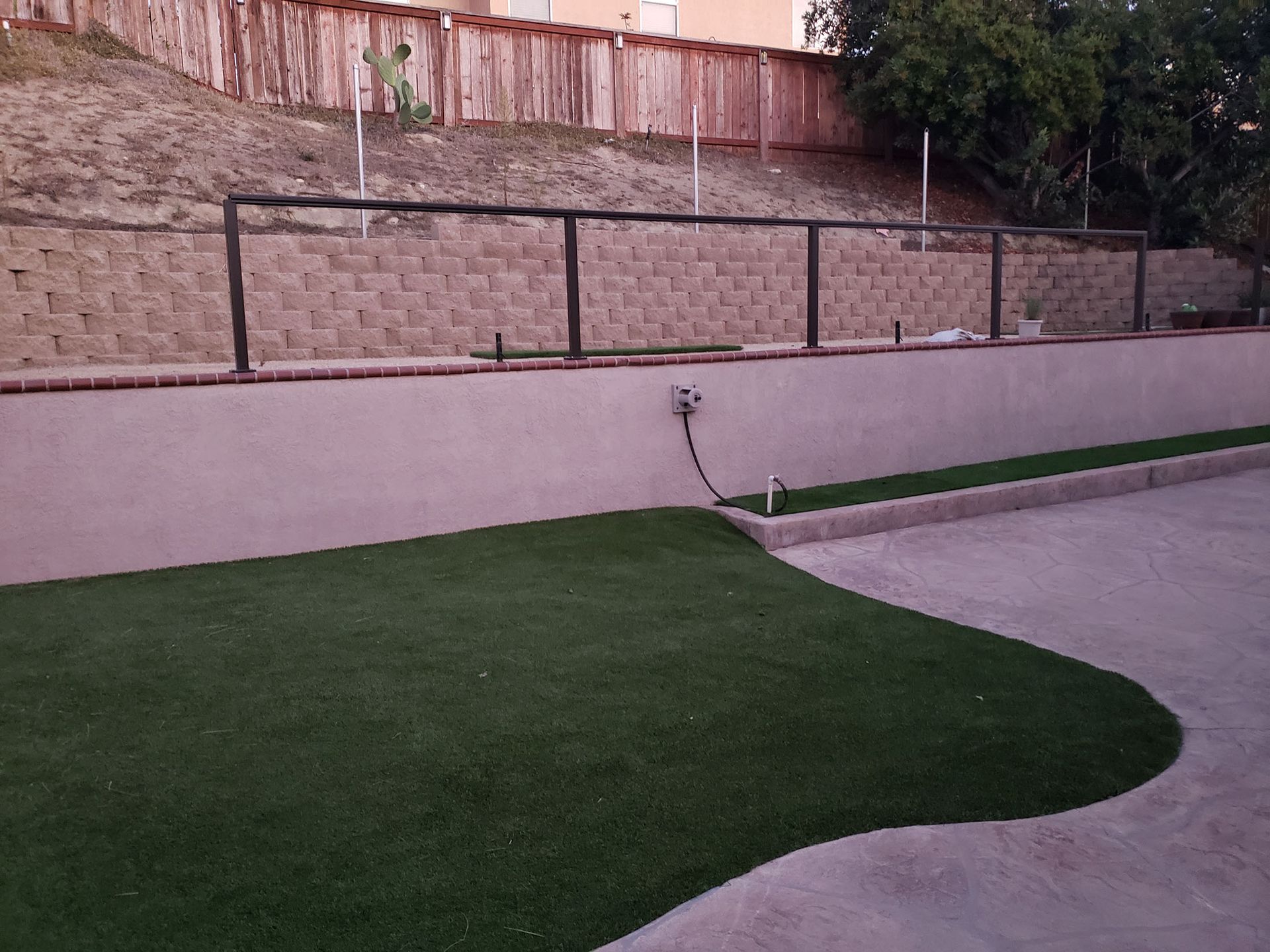 Green artificial turf in a yard, concrete wall with black metal railing, retaining wall, wooden fence, cloudy sky.