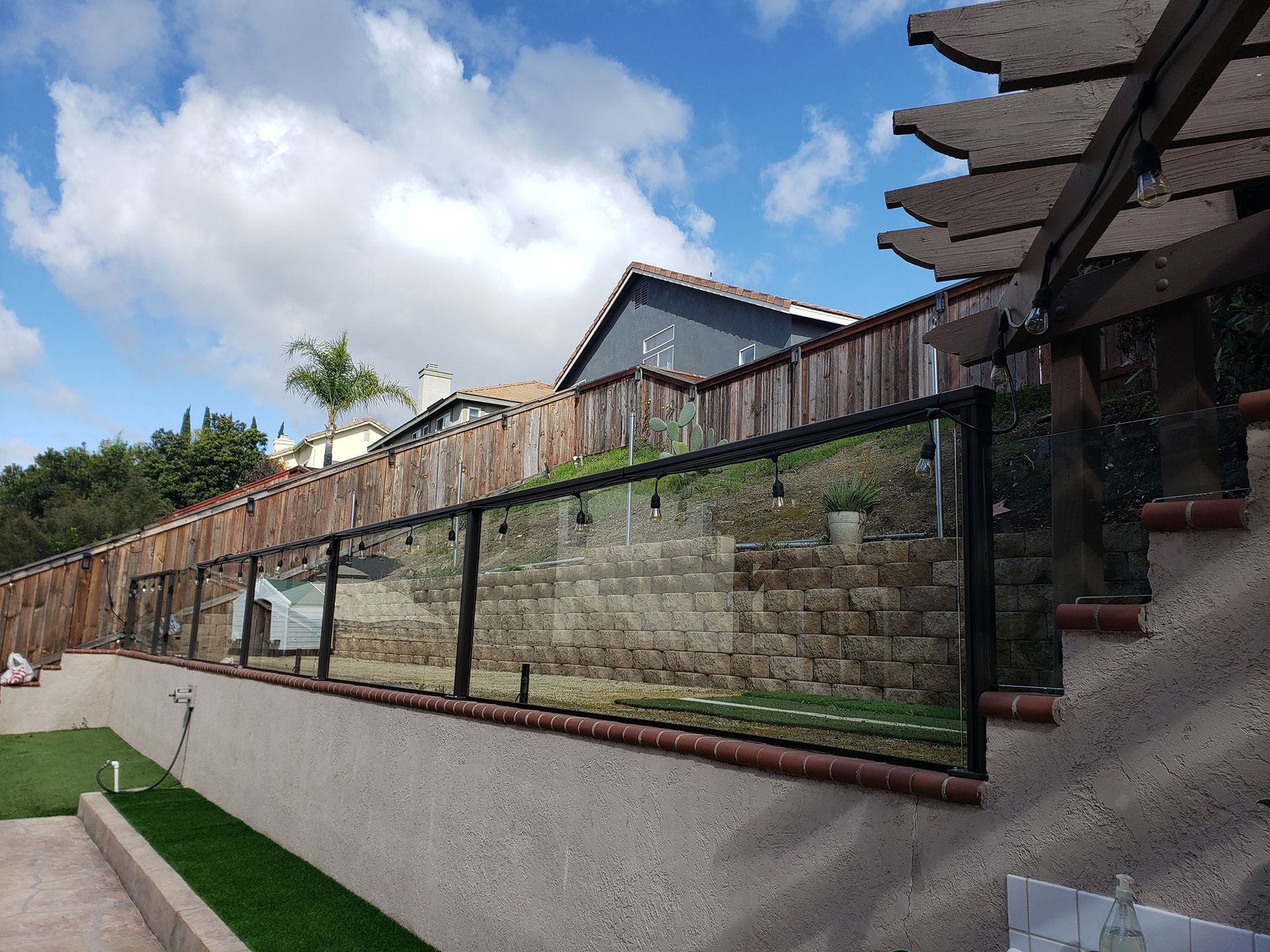 Backyard with glass and wood fences, stone retaining wall, artificial turf, and pergola against a blue sky.