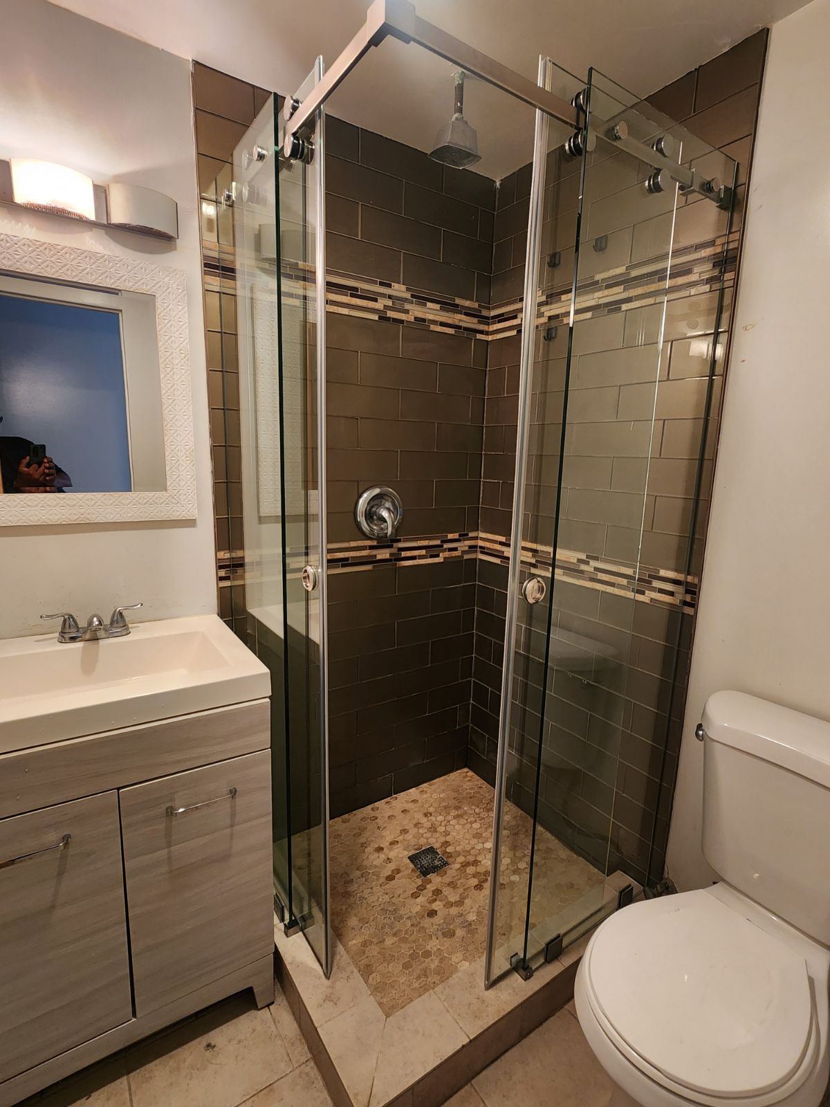 Bathroom with brown-tiled shower, glass doors, and pebble floor. White vanity, toilet, and blue wall.