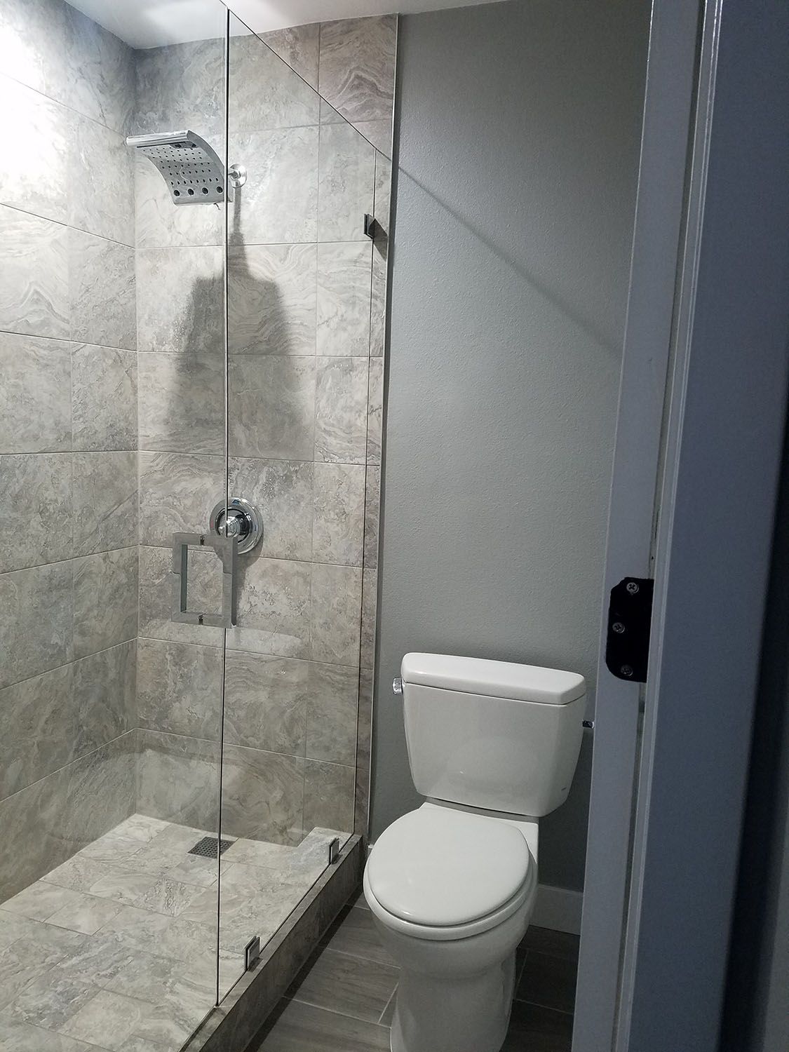 Bathroom with glass shower, gray tiled walls, white toilet, and open door.