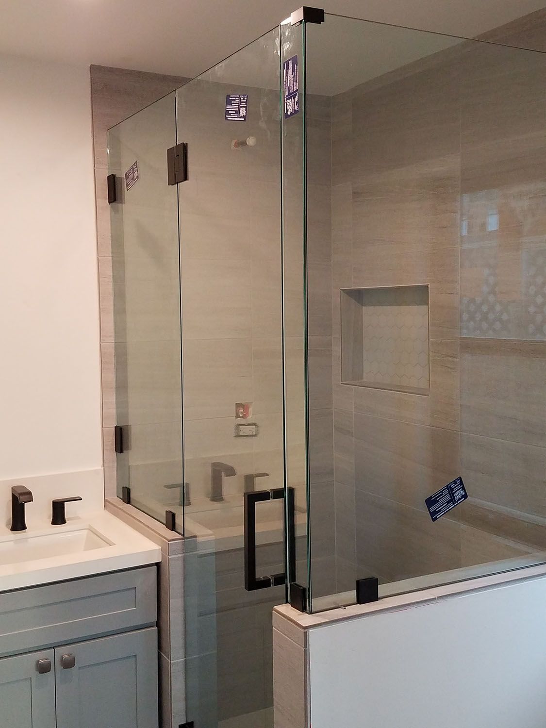Glass shower enclosure with black hardware in a bathroom with neutral-toned tiling and a grey vanity.