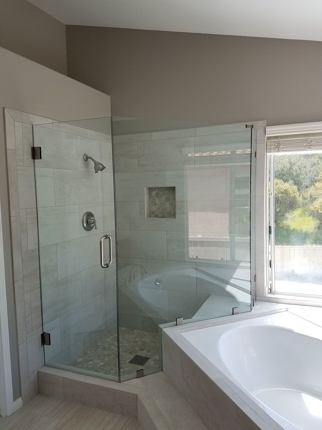 Modern corner shower with glass doors and a built-in bench, next to a bathtub and window.
