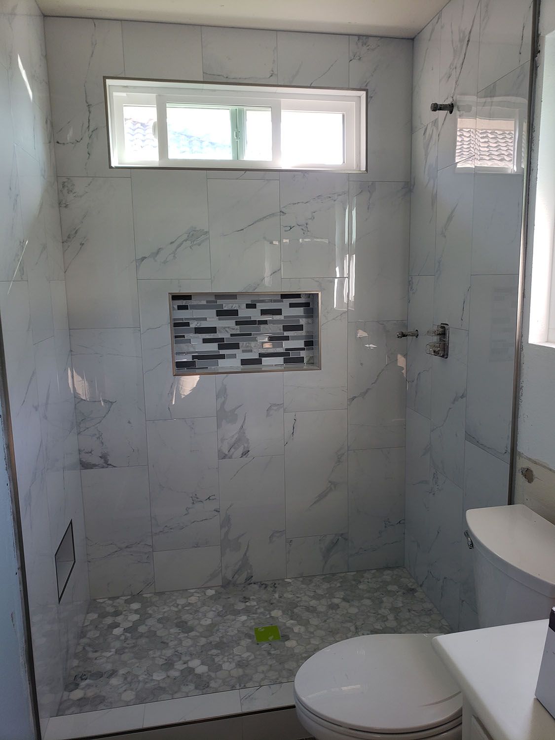 Bathroom with white marble tile, shower, toilet, window, and built-in shelf with decorative mosaic tile.
