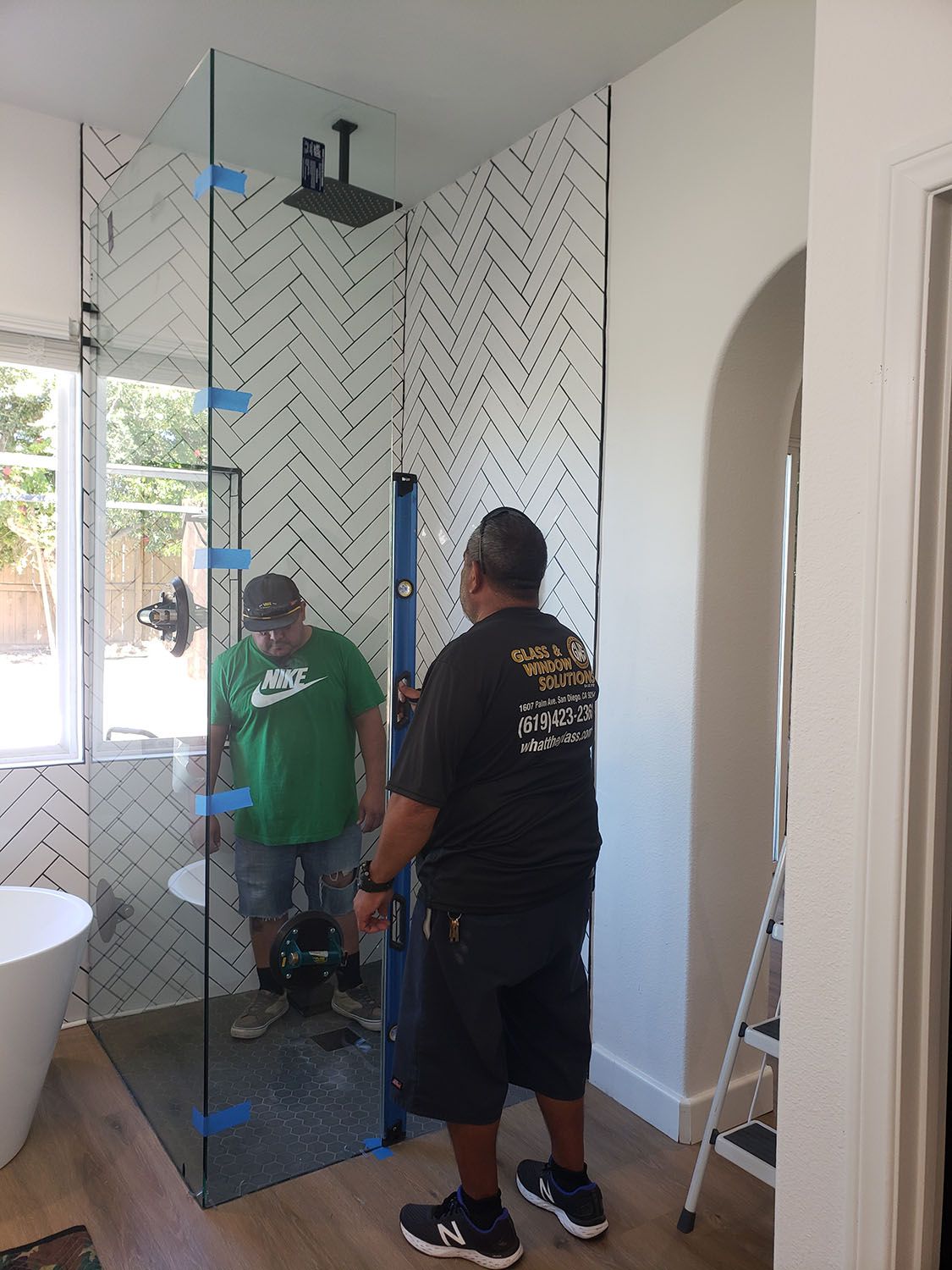 Two men installing a glass shower door in a bathroom with a black and white tiled wall.