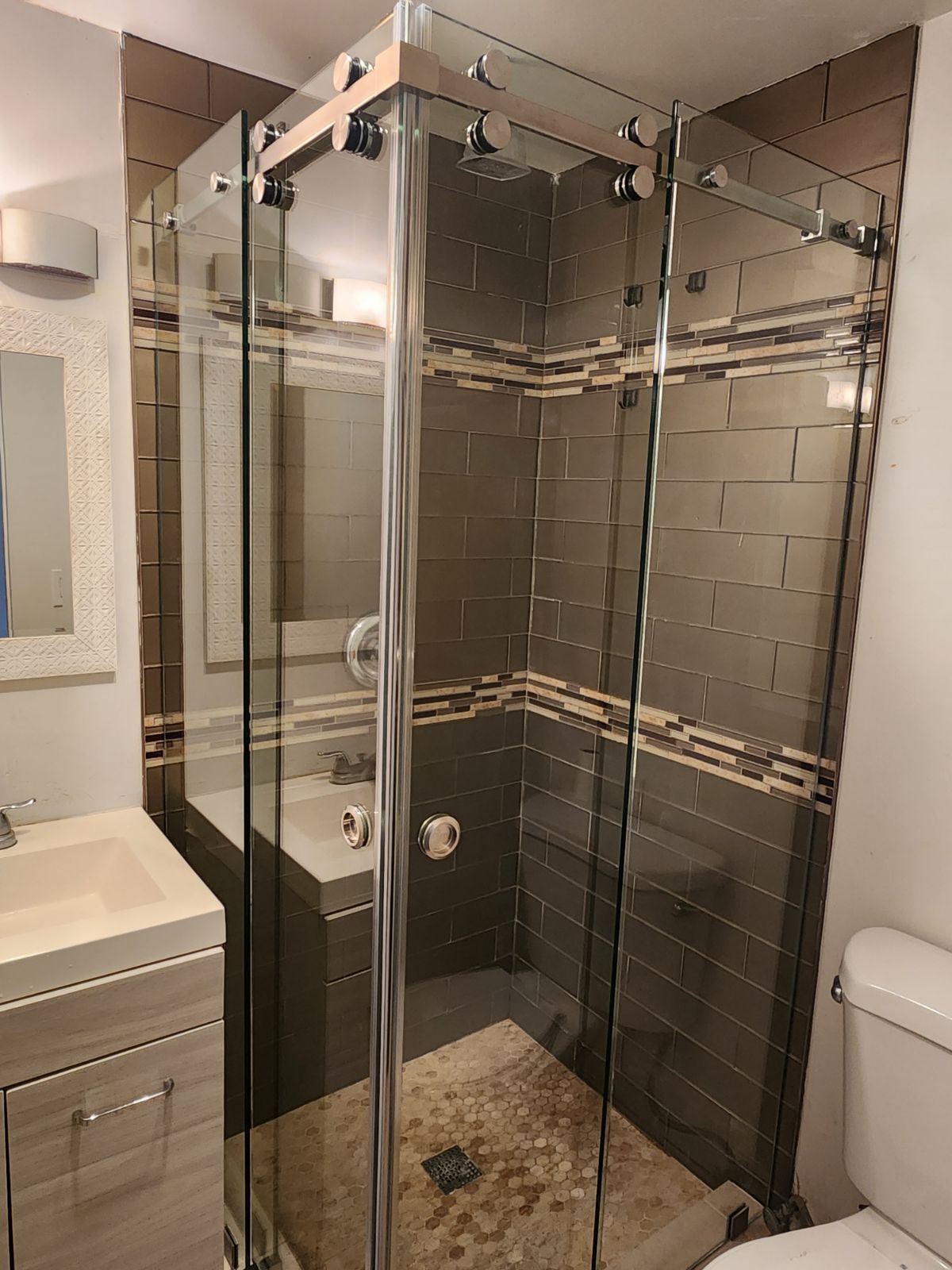 A glass shower enclosure with chrome hardware in a modern bathroom, with dark tile and a vanity.