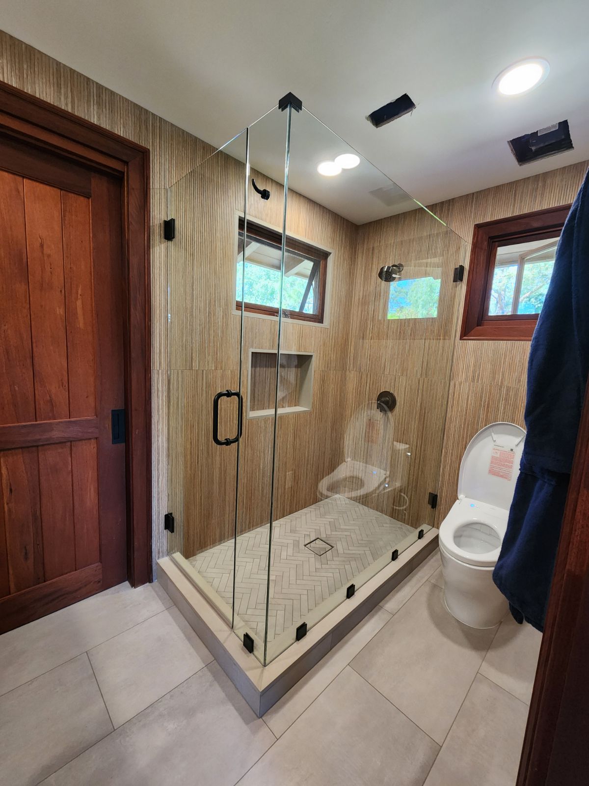 Bathroom with glass shower, wooden door, toilet, and light-colored tiles and walls.
