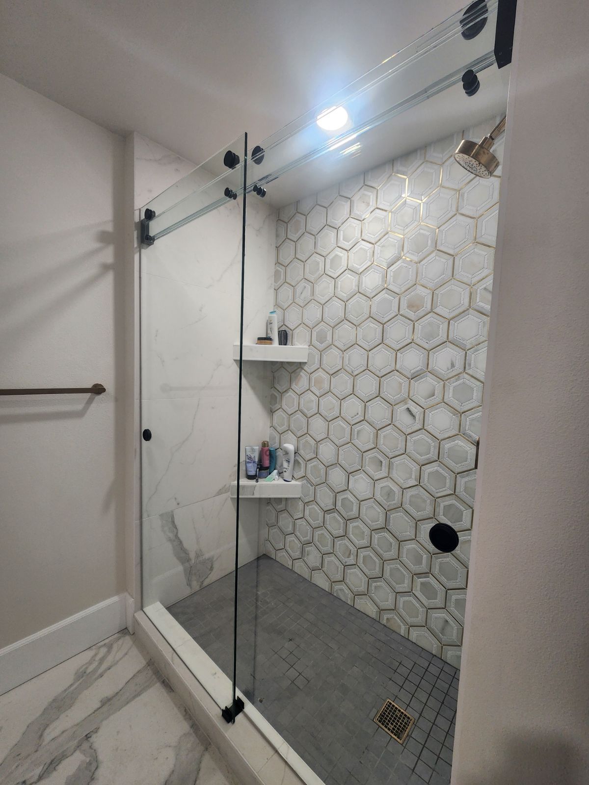 Modern shower with clear glass sliding door, hexagon tile wall, built-in shelves, and gray floor.