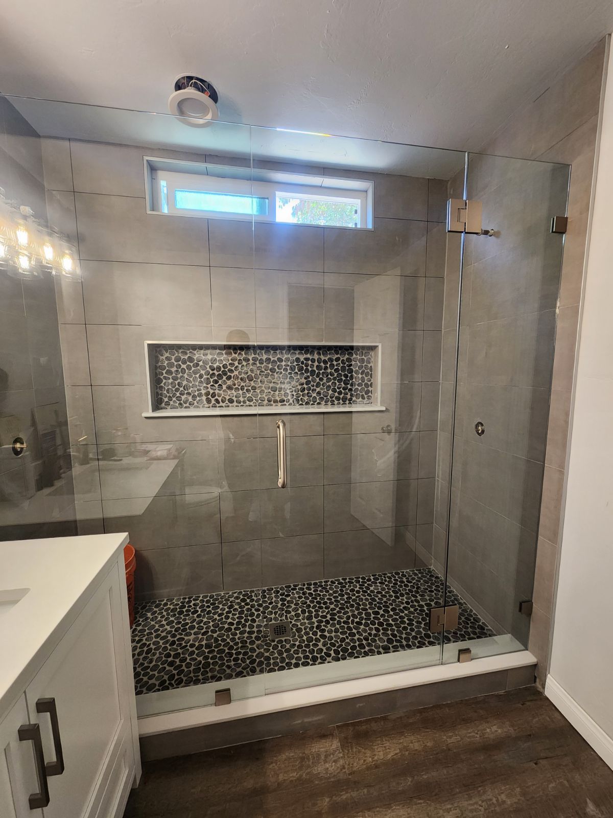 Modern shower with glass doors, pebble floor, built-in shelf, gray tile walls, and a small window.