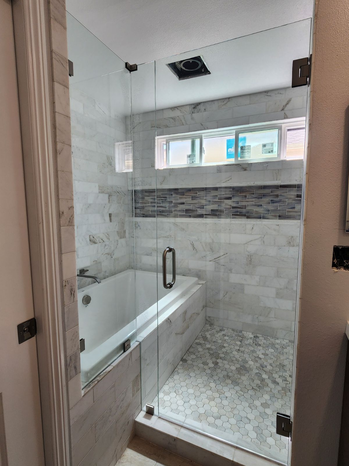 Bathroom with glass shower doors, white tub, marble tile, and pebble flooring.