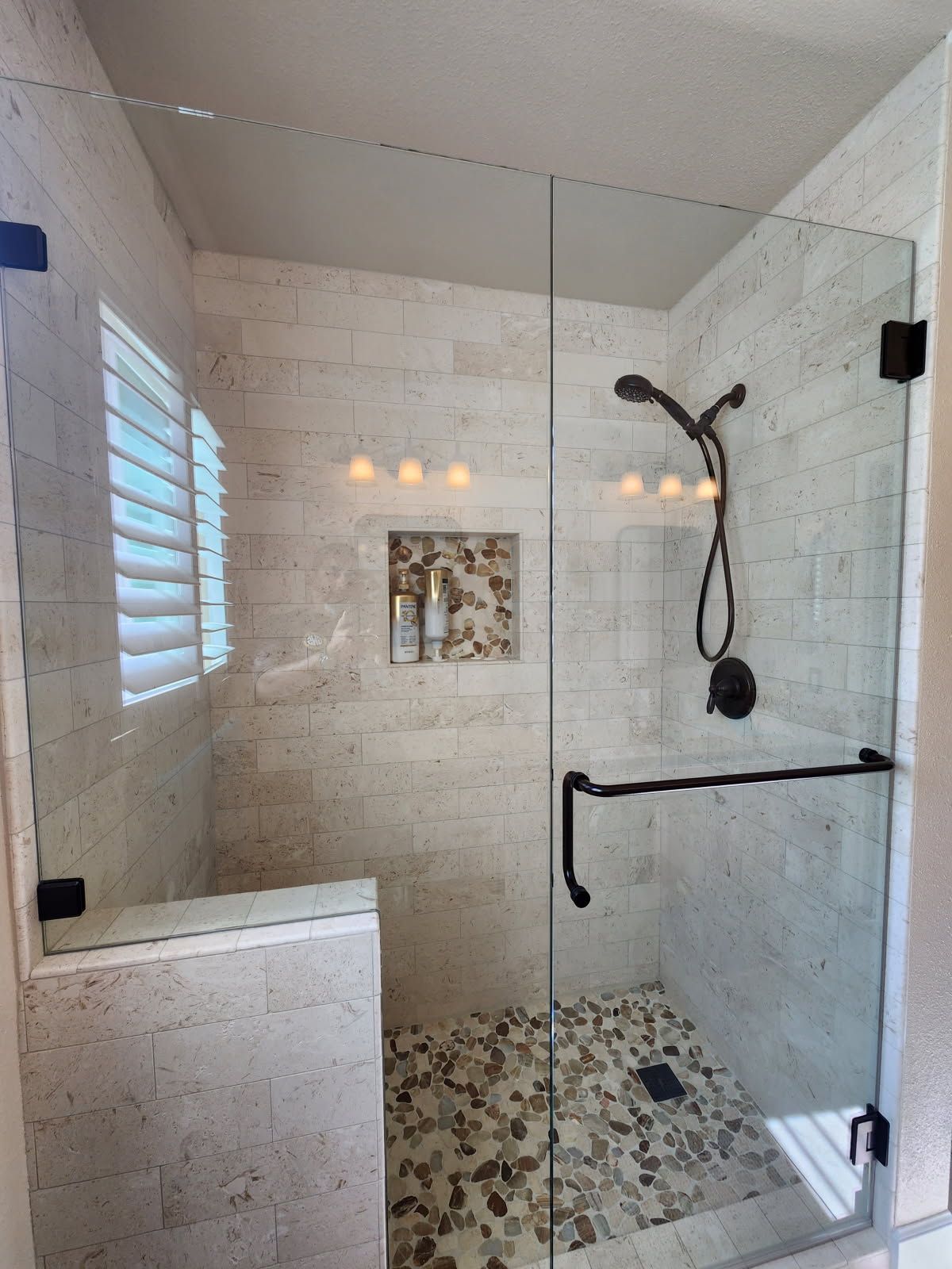 Modern shower with glass door and pebble floor. White brick-like tiles, dark fixtures, and a small window with blinds.
