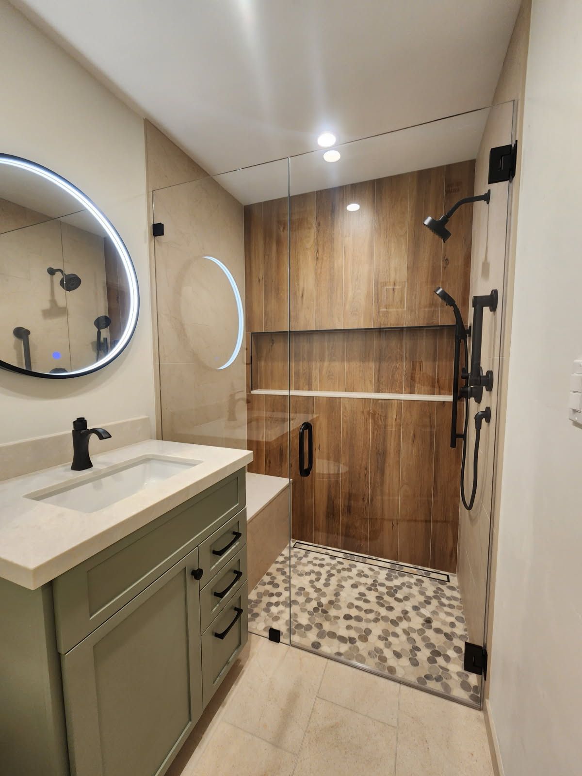 Bathroom with a green vanity, round mirror, and a wood-paneled shower with a glass door and bench.