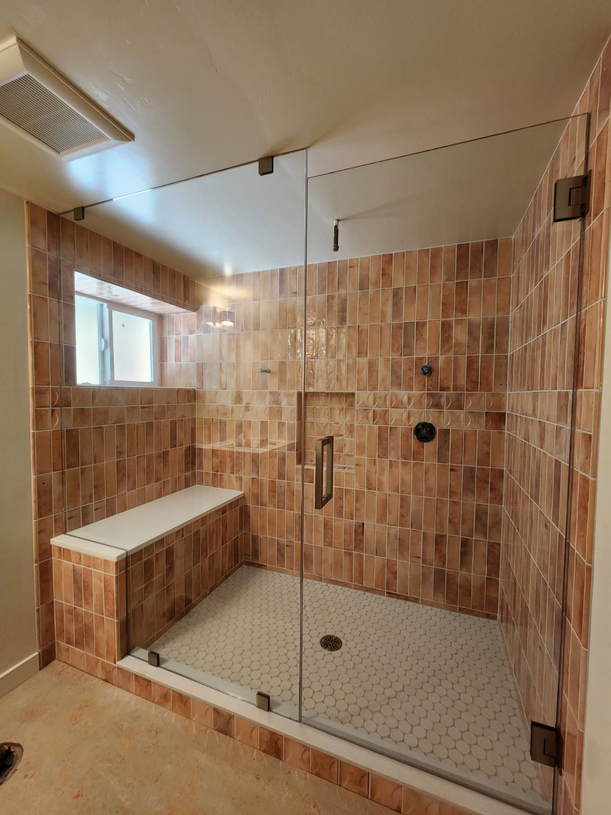 Newly remodeled shower with brown tile walls, a white bench, and glass doors. White honeycomb tile floor.
