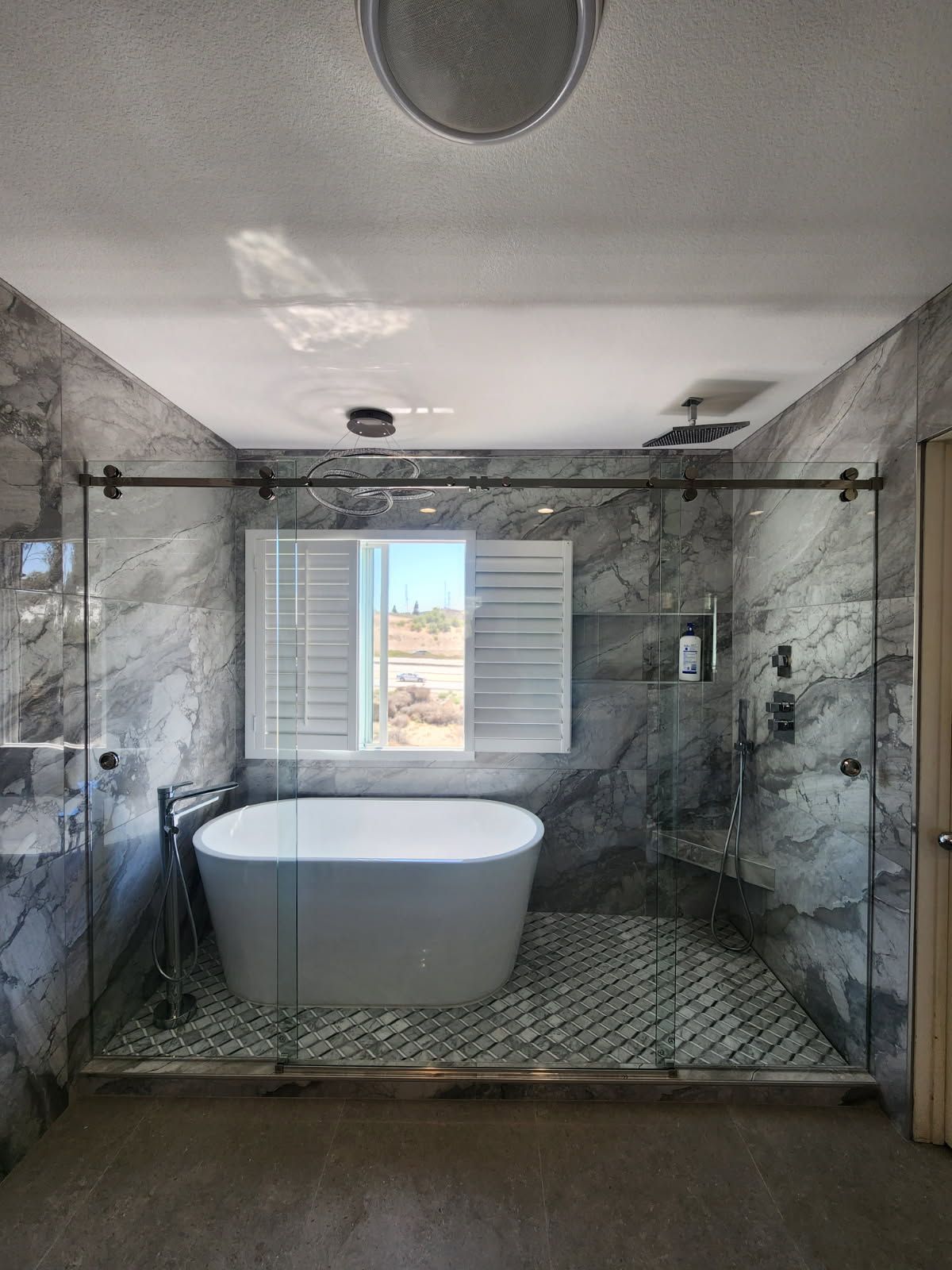 Bathroom with marble walls, glass shower enclosure, freestanding tub, and a window with closed shutters.