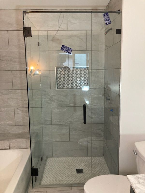 Glass shower door with dark hardware, gray tiled walls and floor, next to a white toilet.