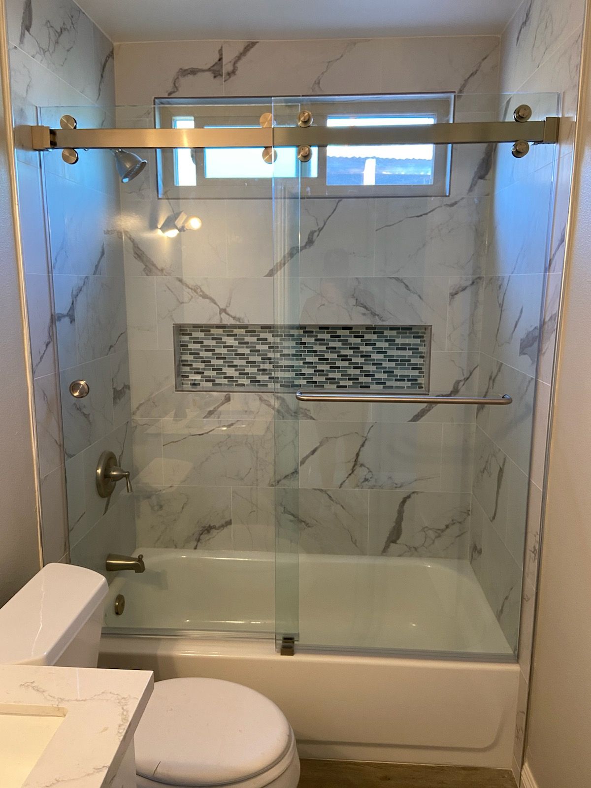 Bathroom with marble-look tile, glass shower door, and a built-in shelf with mosaic tile. A toilet is visible.