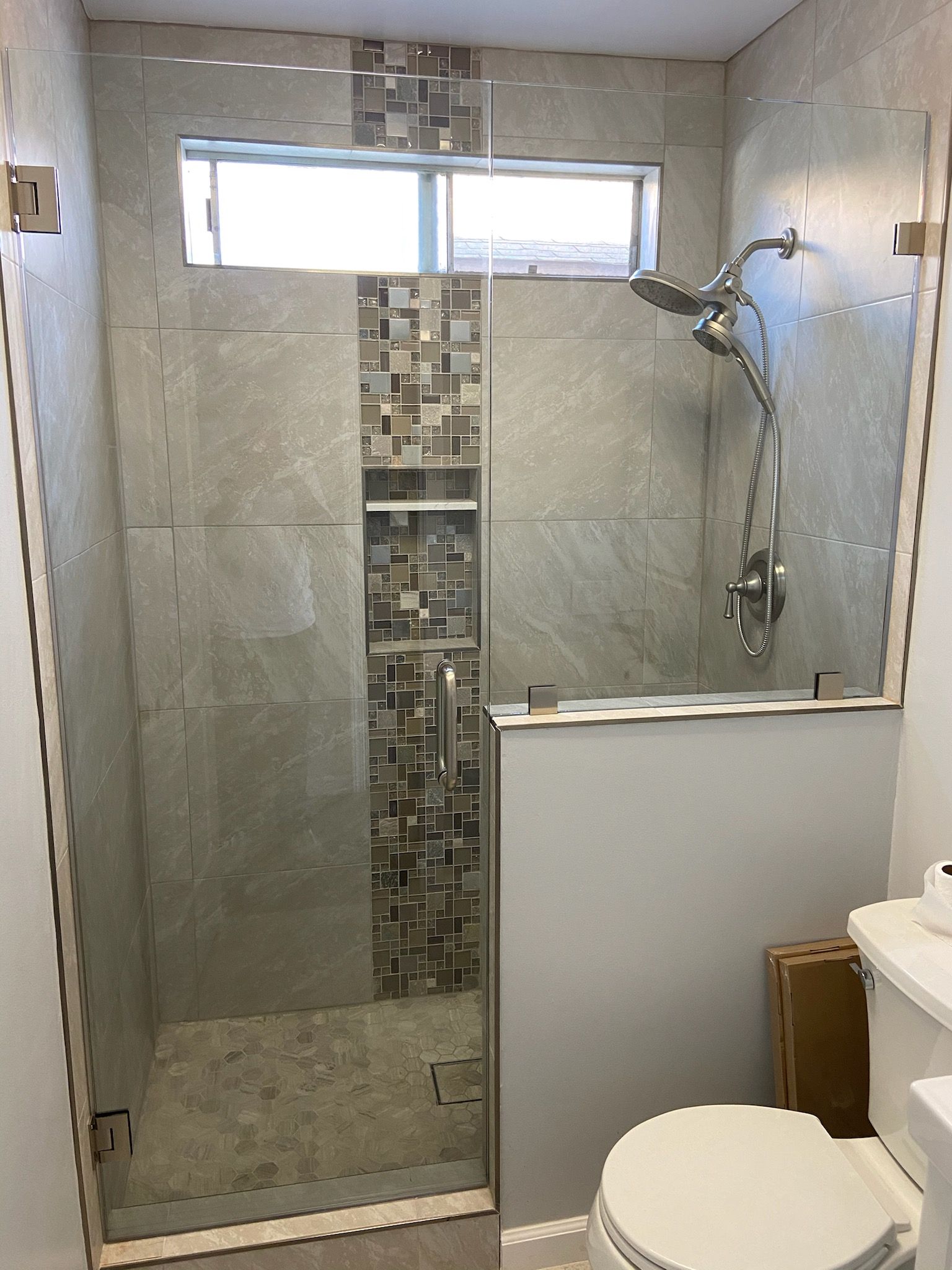 Bathroom shower with gray tiled walls, glass doors, and a decorative mosaic strip.