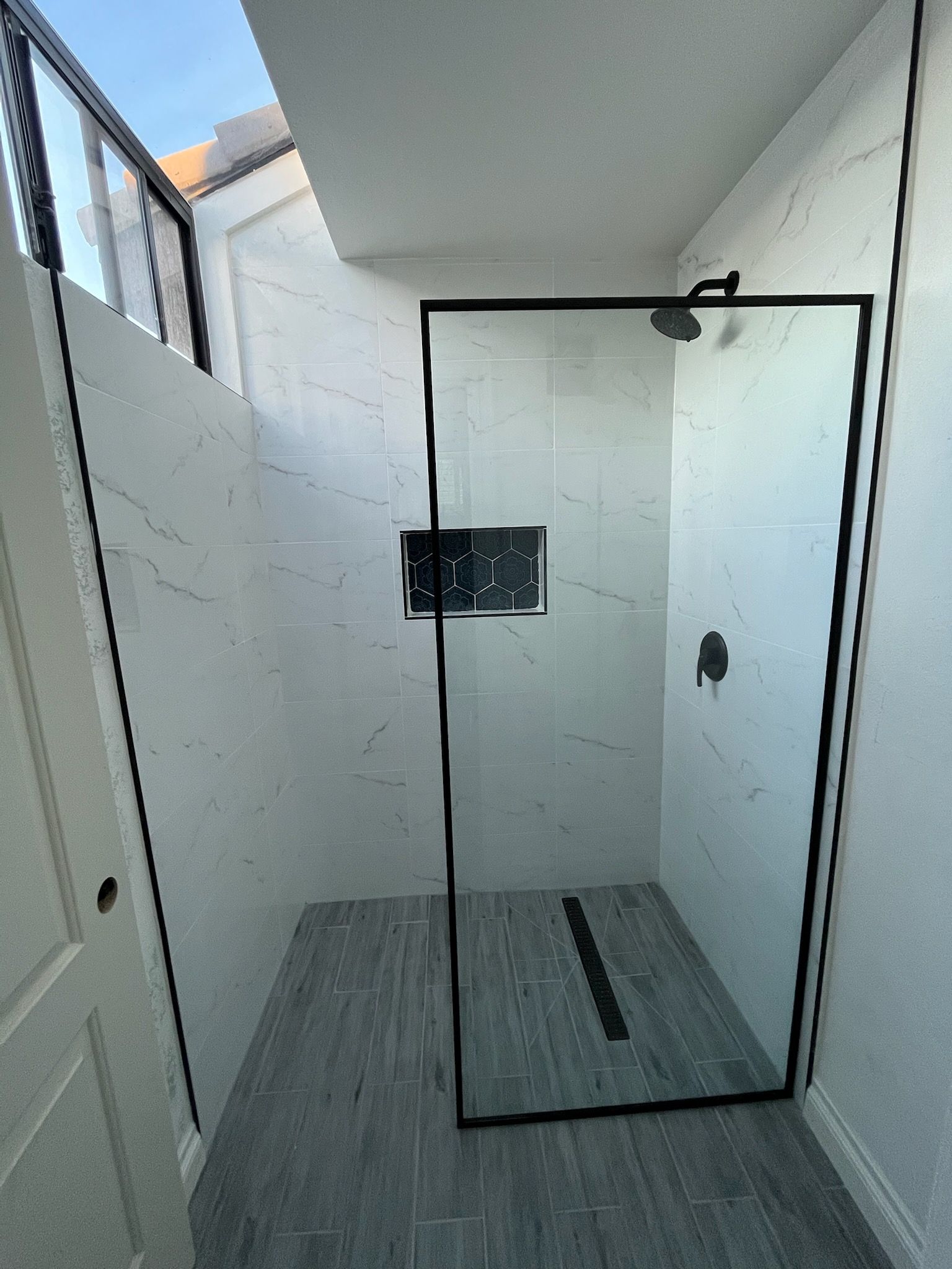 Shower with a black-framed glass door, white tiled walls, gray floor, and a black showerhead.