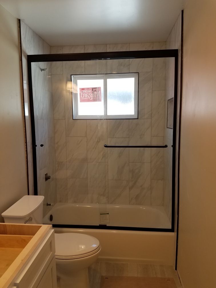 Bathroom with white tub, toilet, and marble-like tile. Glass shower door with dark frame. Window with sticker.