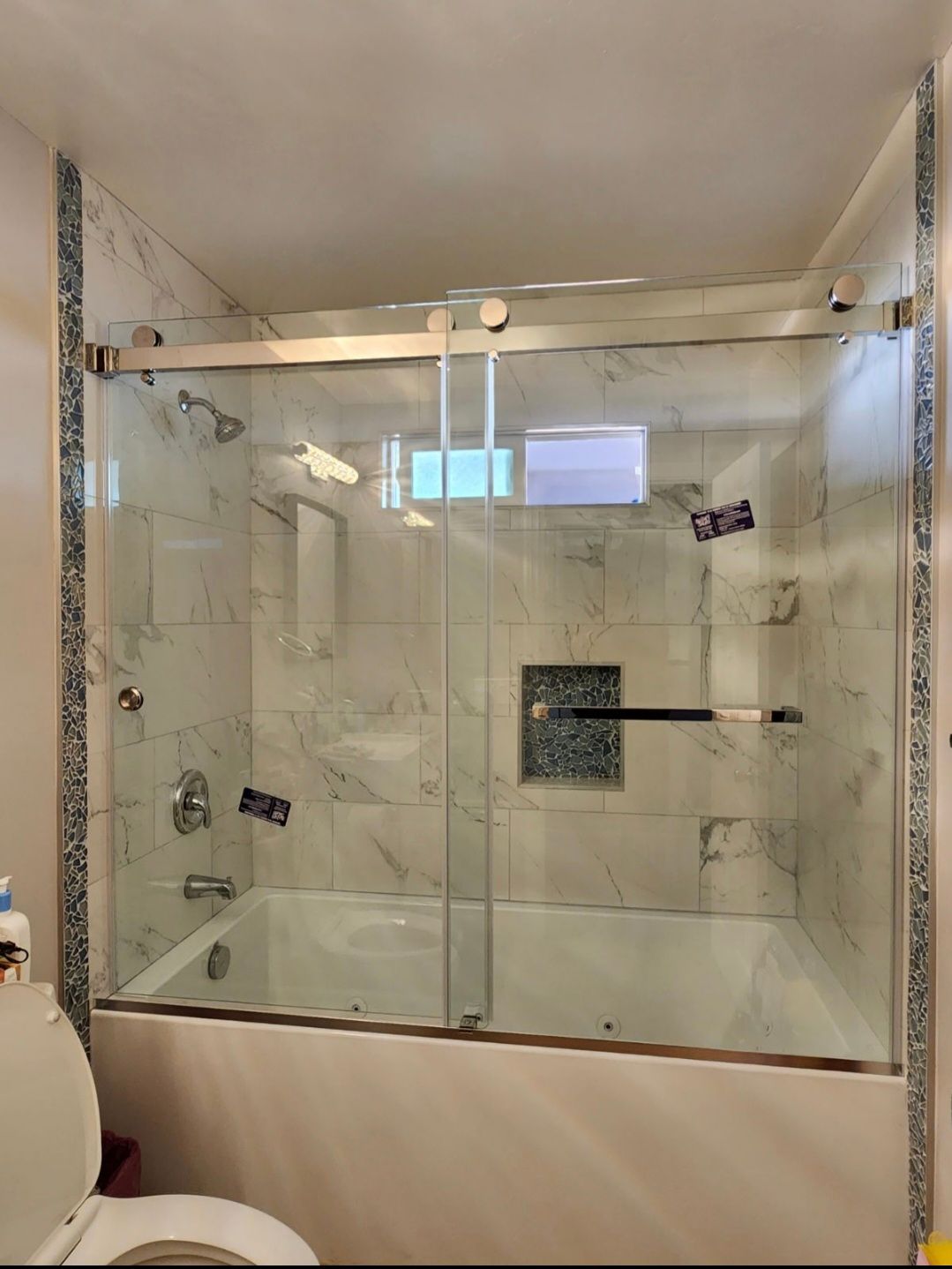 A clear glass shower door with gold hardware, over a white bathtub with marble tile walls.