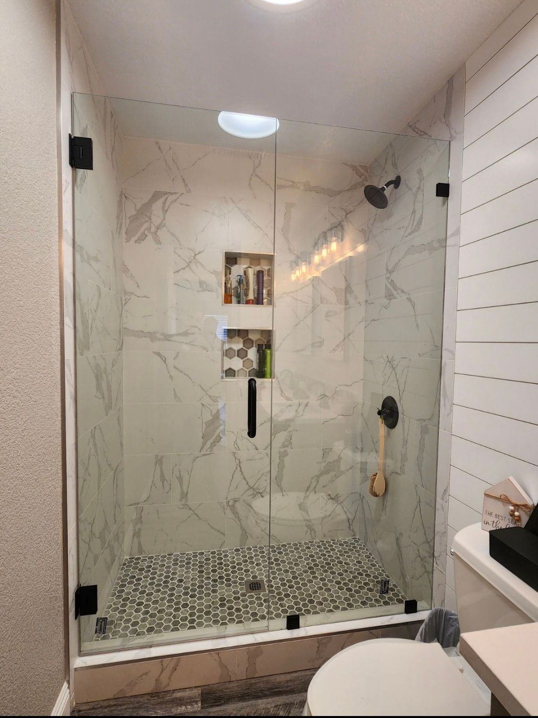 Shower stall with marble-like tile, pebble floor, and glass doors with black hardware.