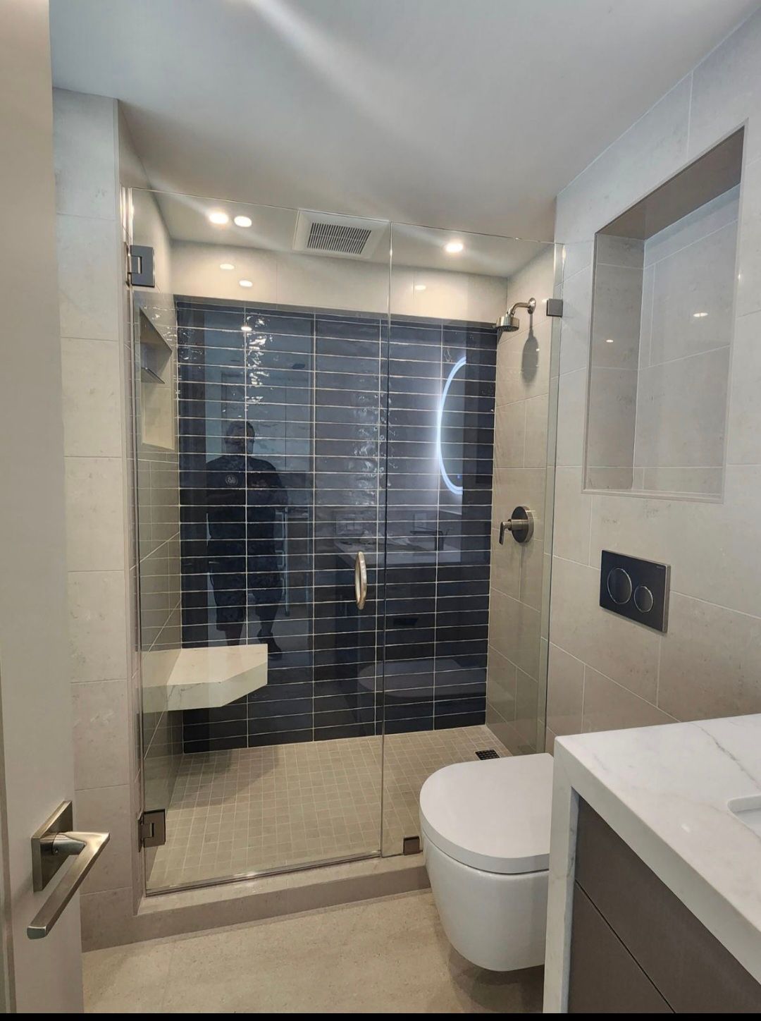 Modern bathroom with a glass shower, floating bench, and toilet. Beige and dark tile, chrome fixtures.