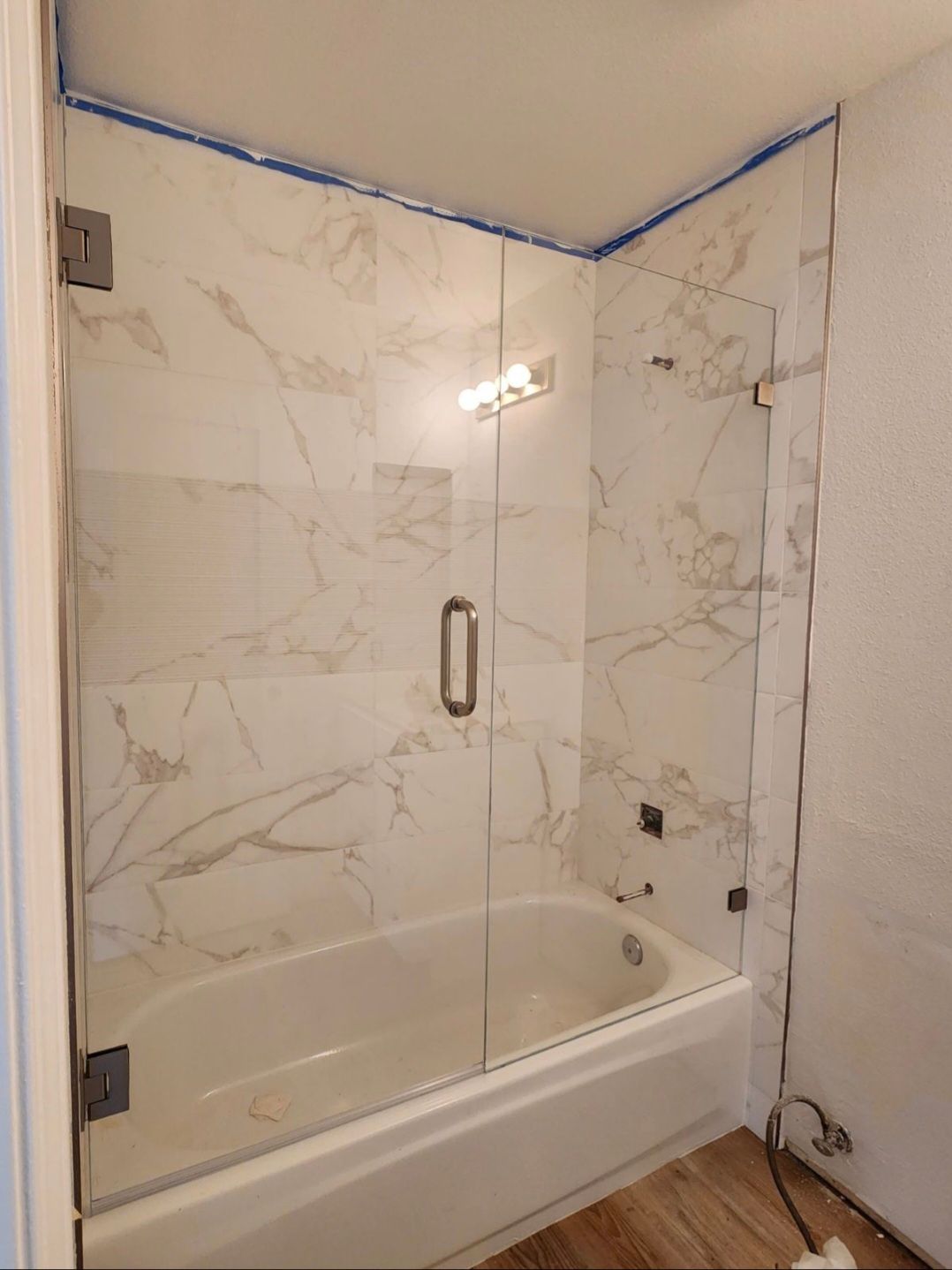 A glass shower enclosure with a door over a white bathtub. The walls are tiled in marble.