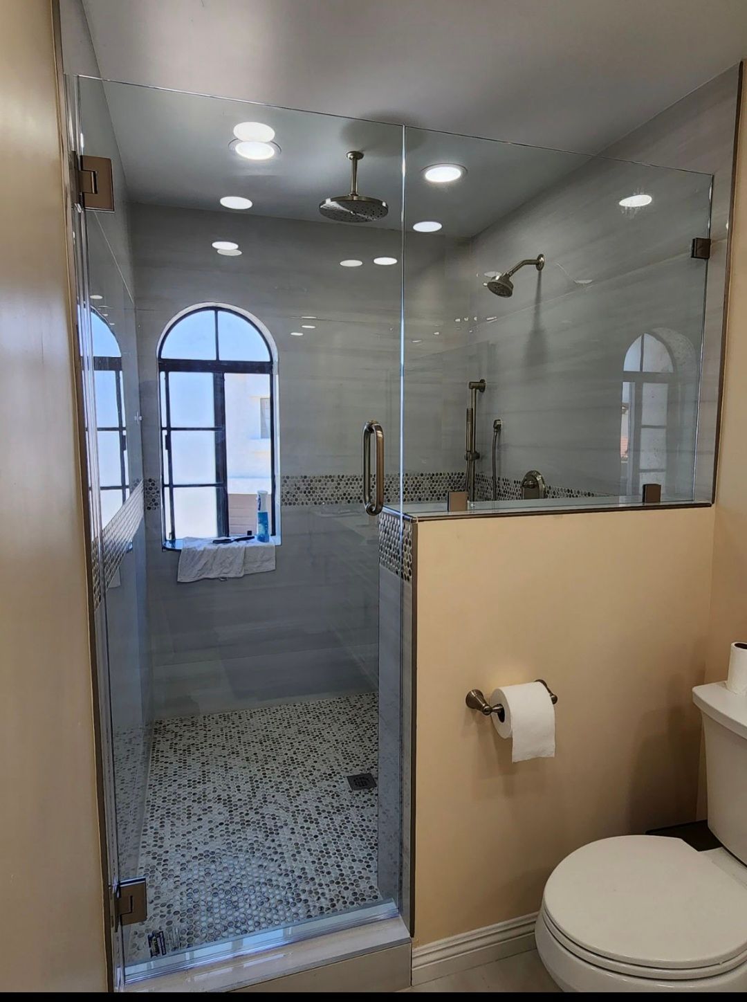 Shower with glass enclosure, toilet, and arched window. Beige walls, light-colored tile, and pebble shower floor.
