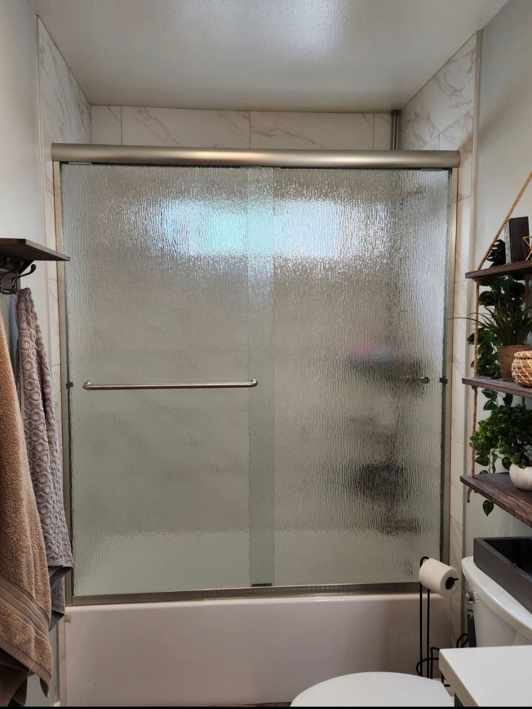 A shower with a textured glass door, silver frame, and white tile. A towel rack with a towel is to the left.