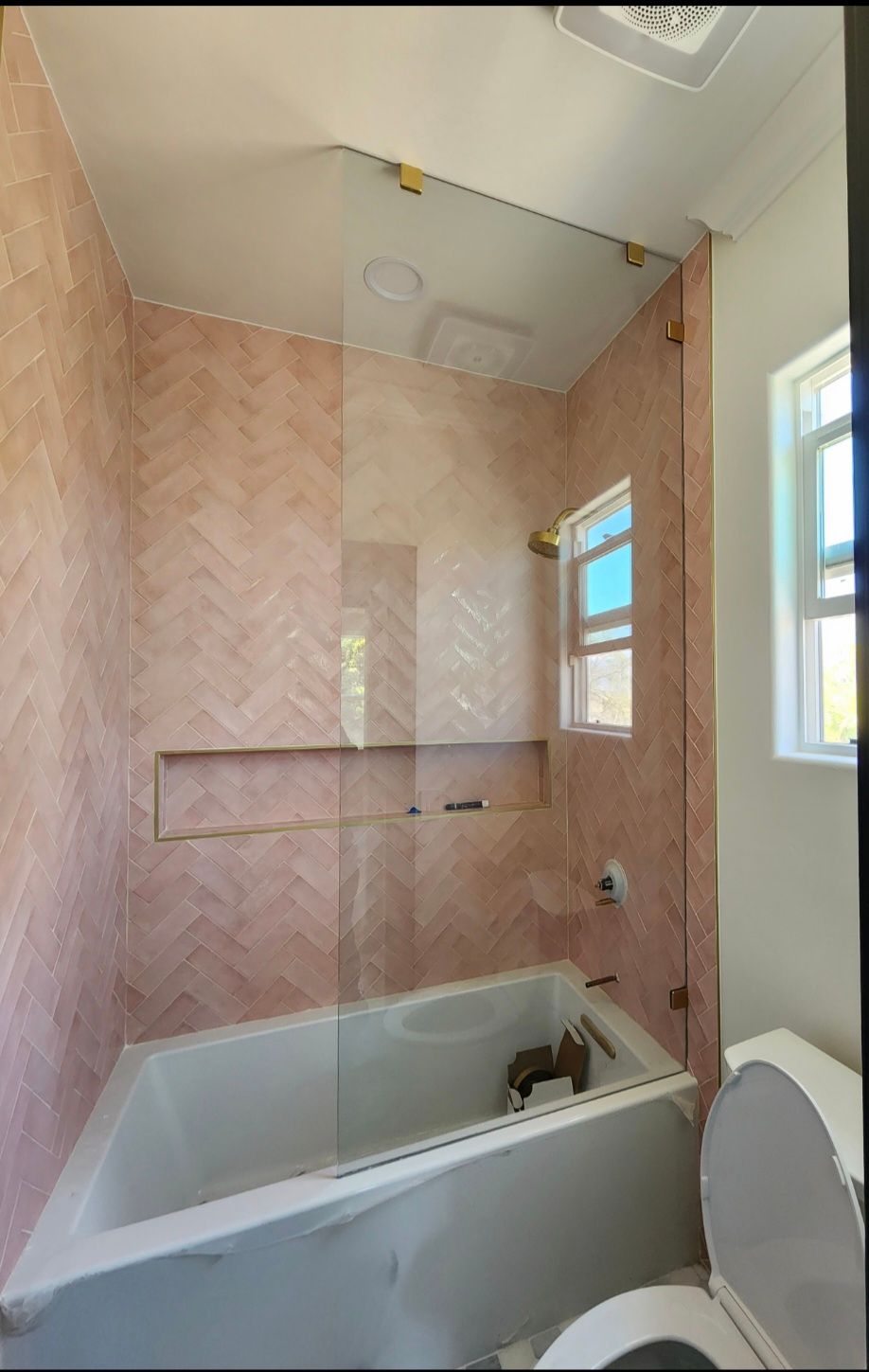 Pink-tiled bathroom with a bathtub, glass shower panel, toilet, and small window.
