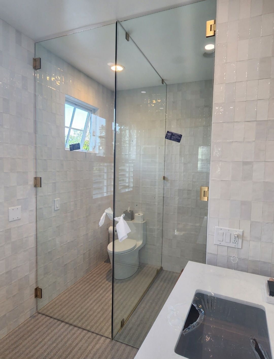 Glass shower enclosure in a bathroom with a toilet and window. Beige tile walls.