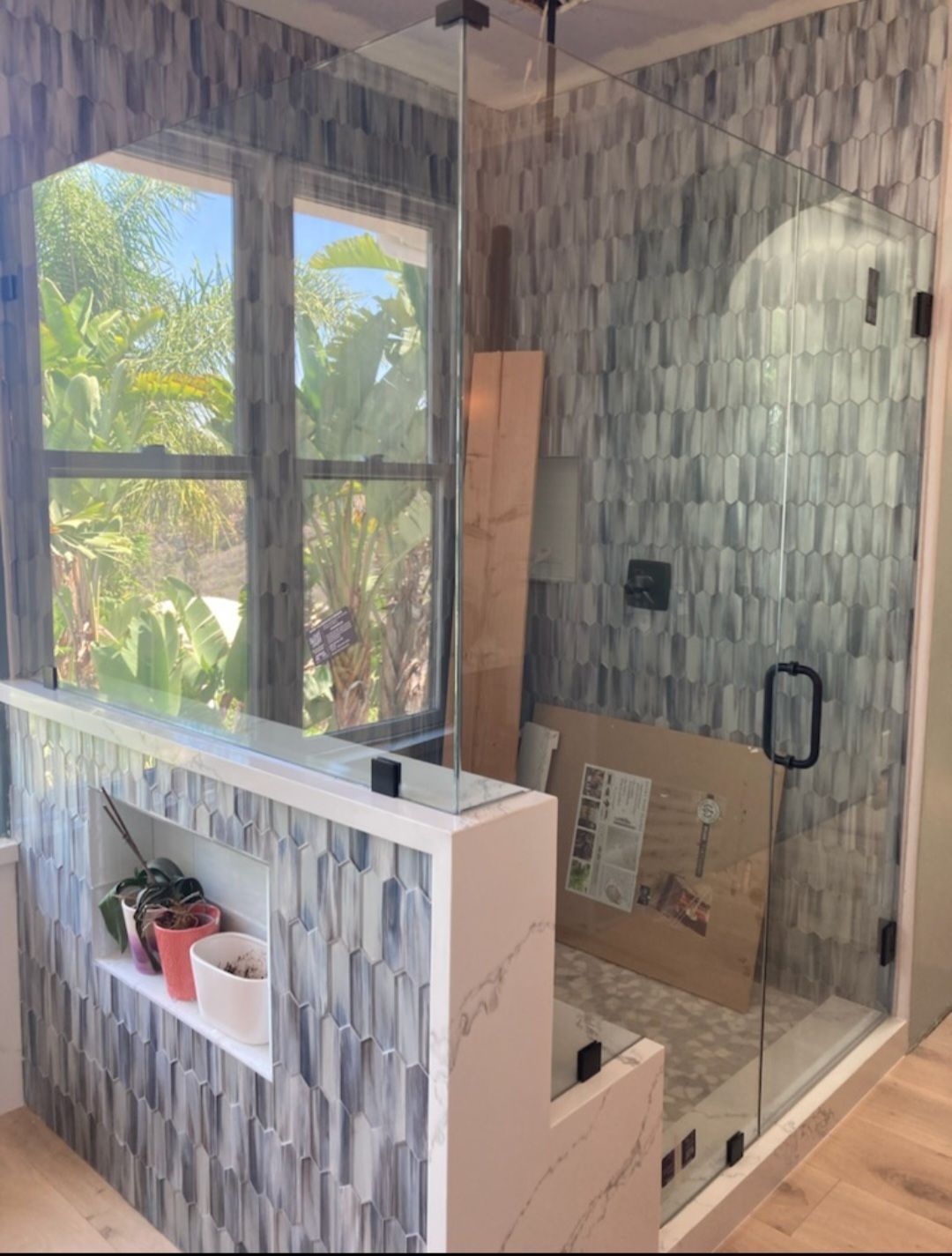 Shower with glass door, tiled walls, built-in shelf, and window overlooking greenery.