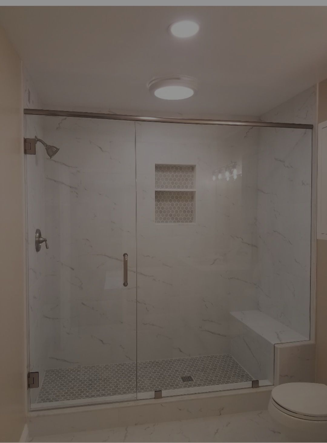 Glass shower enclosure with white marble tiles and built-in bench. Recessed shelf and circular ceiling light.