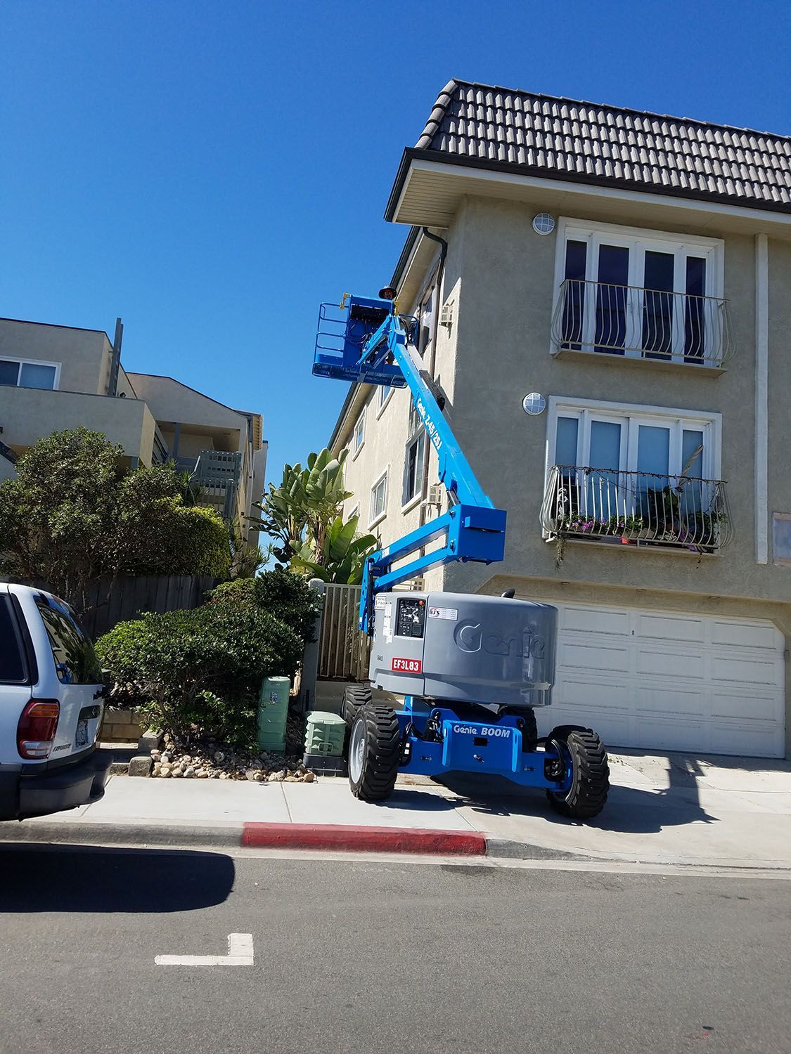 A blue boom lift extending towards a light-colored building. The lift is parked on a street.