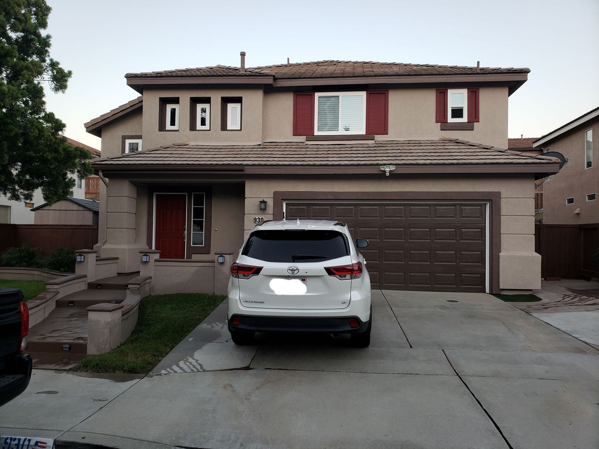 Two-story tan house with a white SUV parked in the driveway. Red accents on the house, brown garage door.