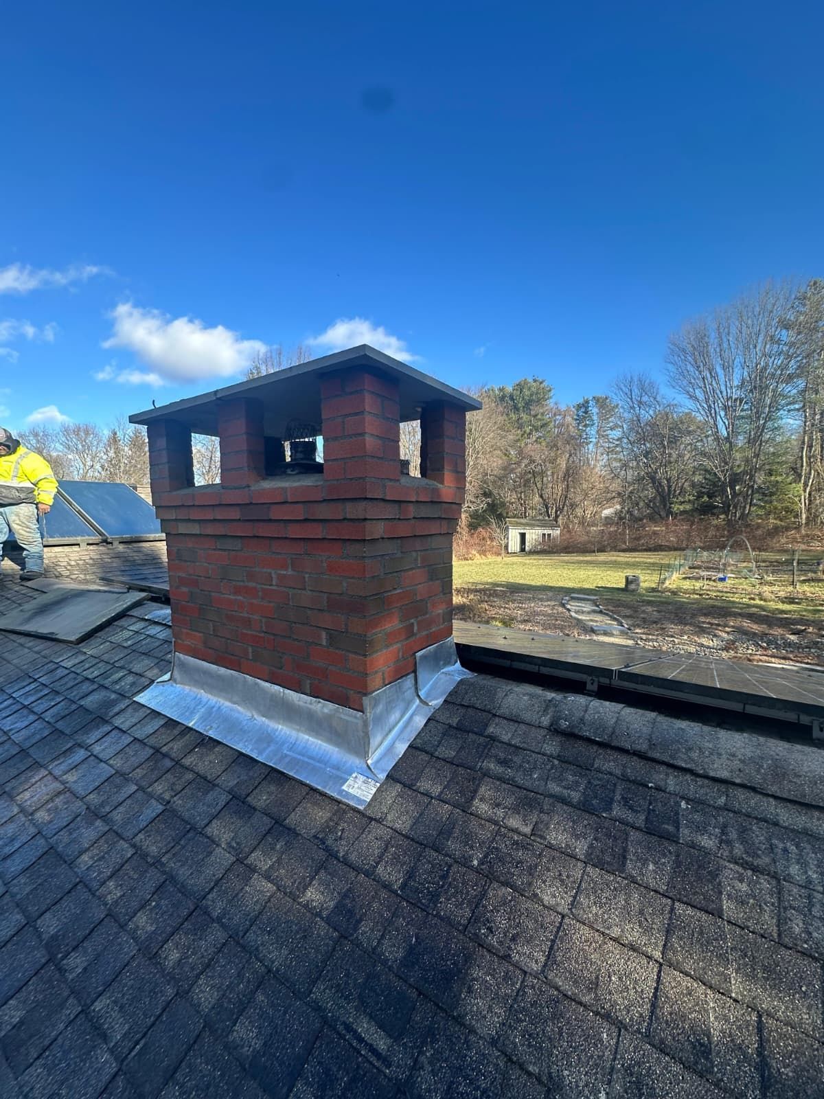 Brick chimney on a shingled roof under a blue sky, with a worker in a yellow jacket nearby.