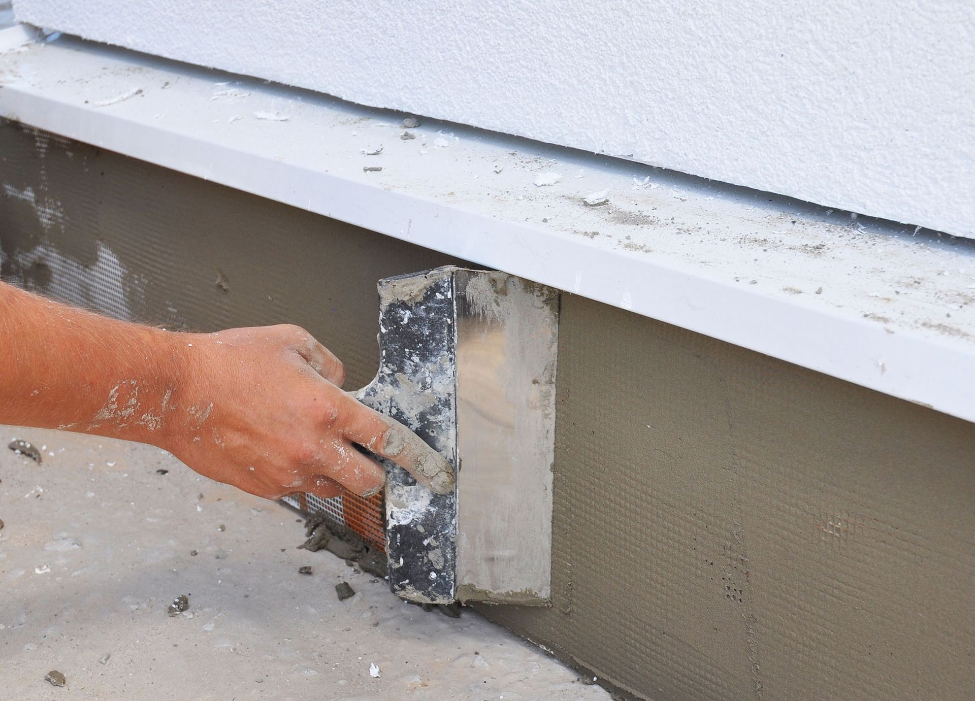 Hand using a trowel to apply gray plaster to the base of a white wall.