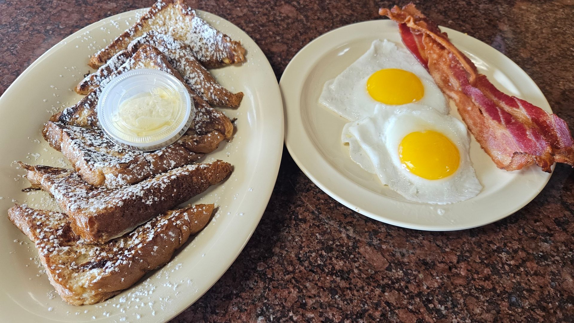 A breakfast plate of French toast with butter, next to a plate with two sunny-side-up eggs and a strip of bacon.