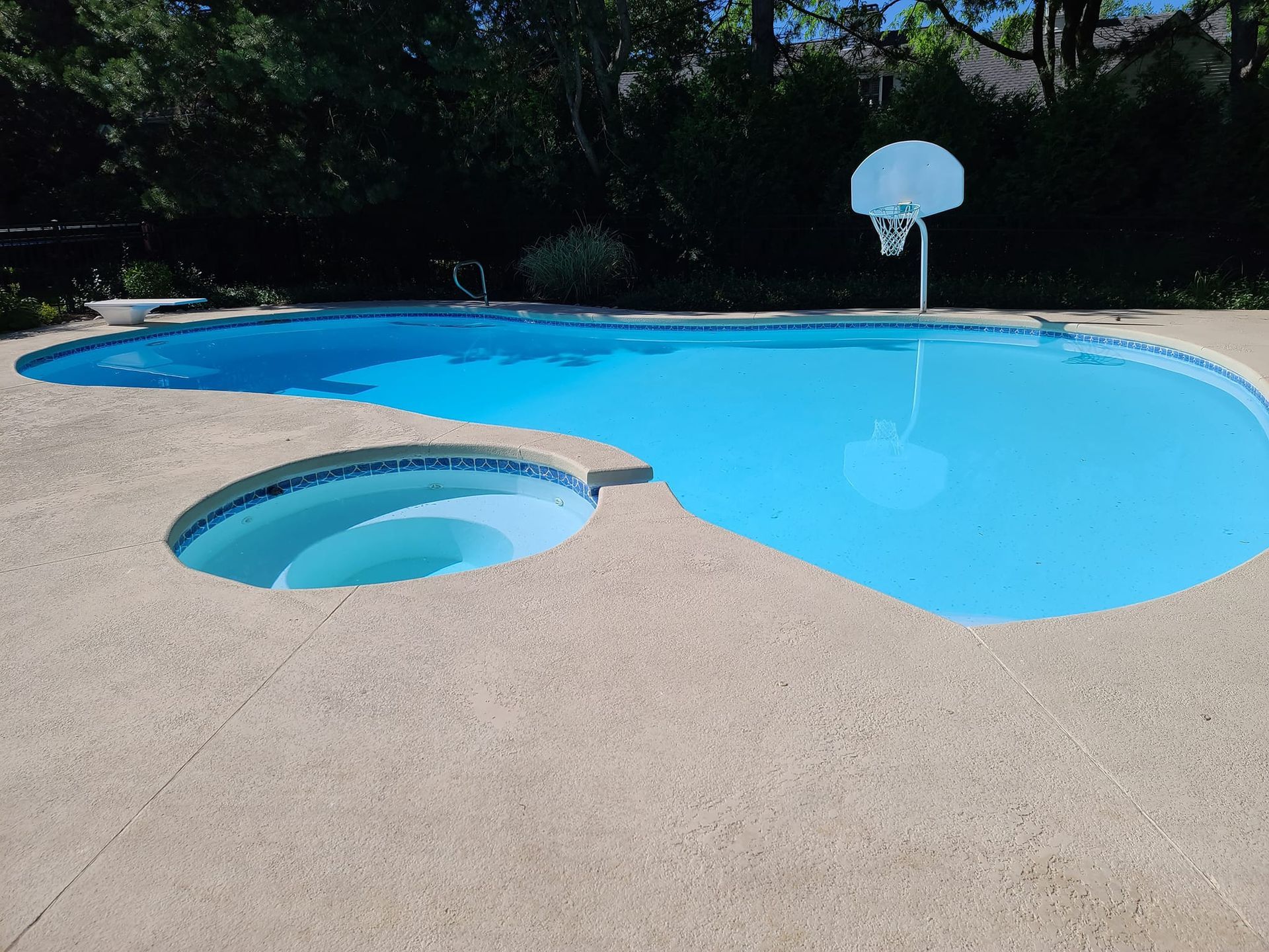 A kidney-shaped swimming pool with an attached circular spa and a basketball hoop, set on a textured concrete patio.
