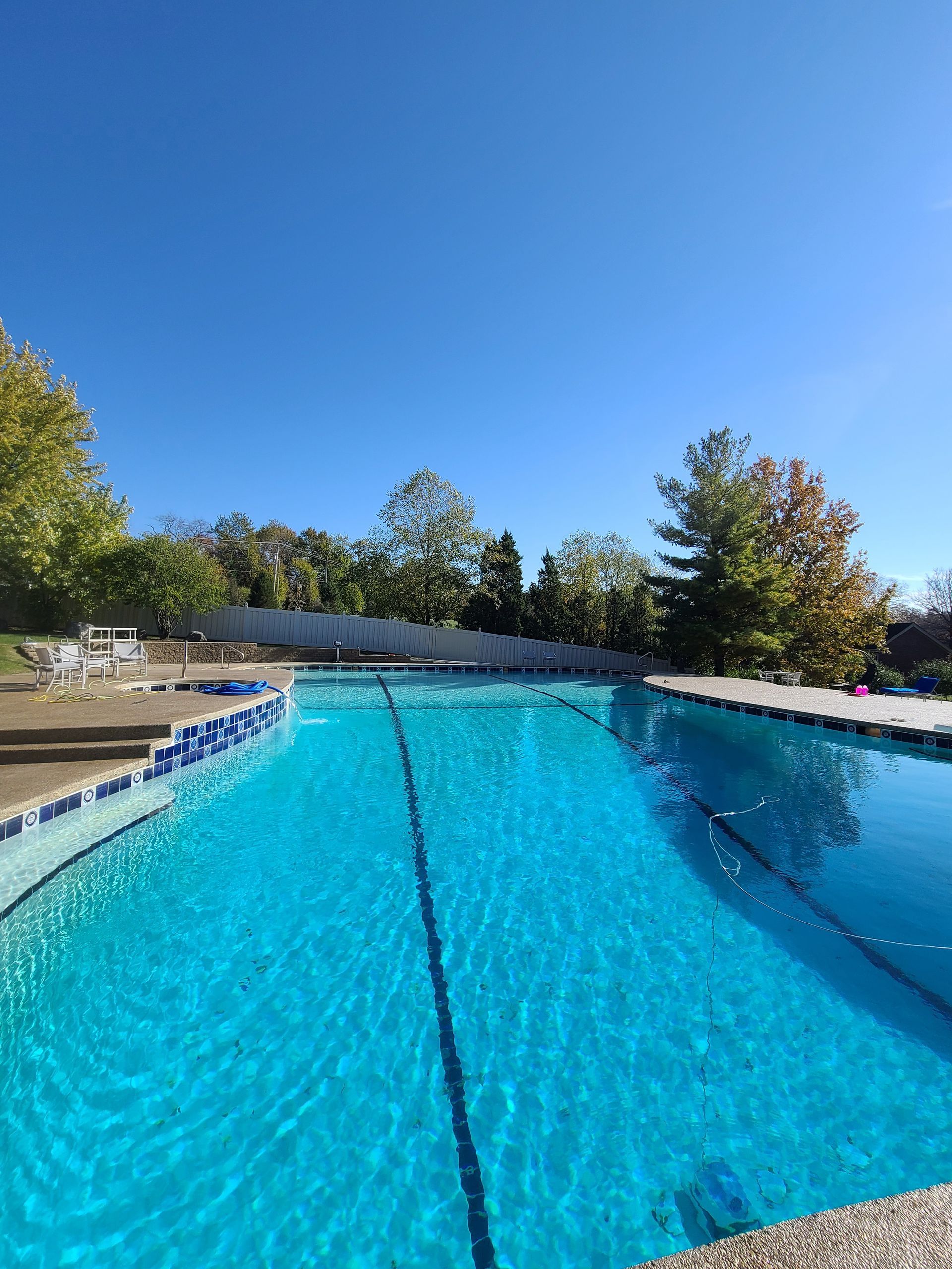 An outdoor swimming pool with blue water and lane markers, surrounded by trees under a clear, bright blue sky.